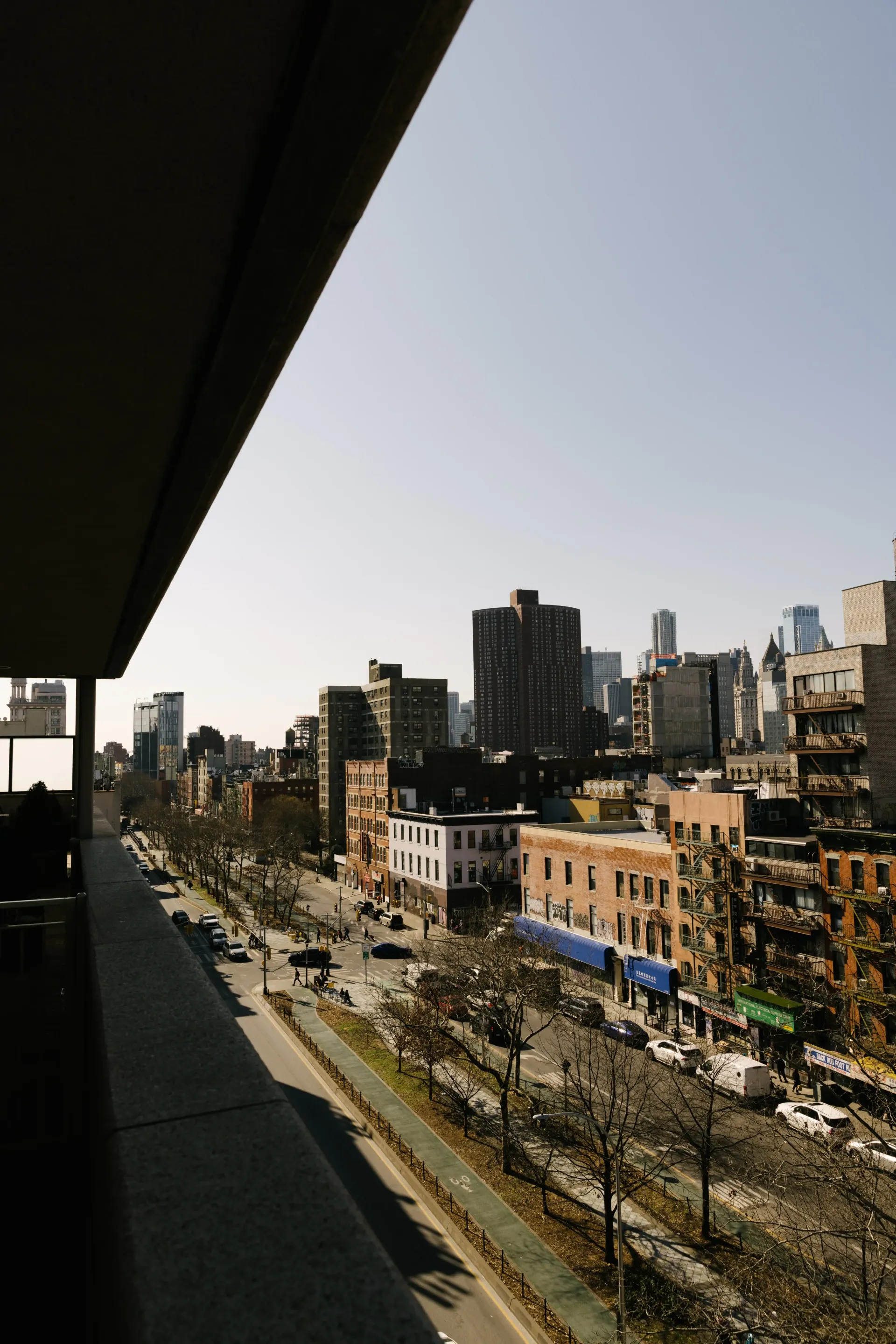 Cityscape viewed from a balcony on a sunny day. Buildings and street with sparse trees.