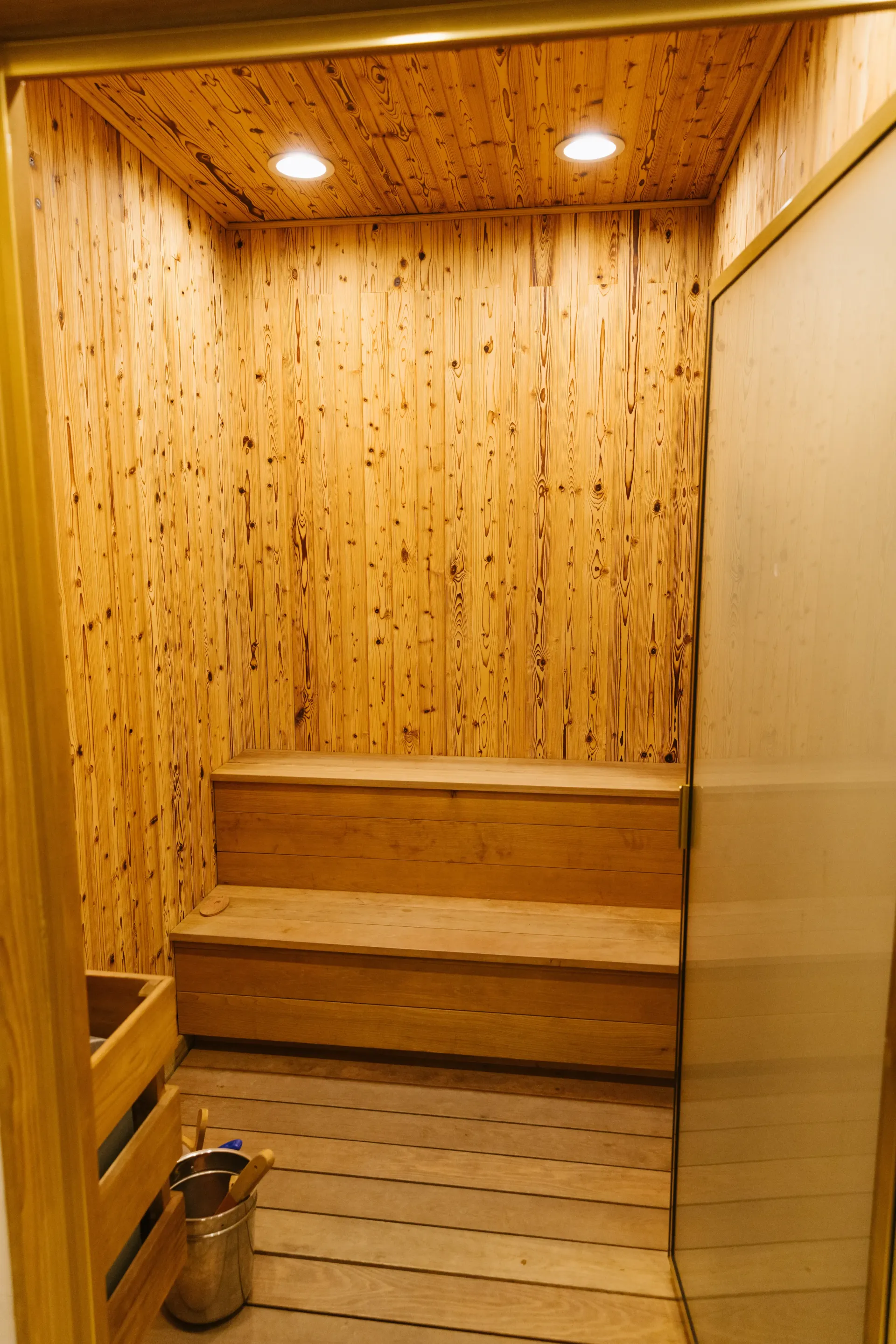 Interior of a wooden sauna with benches and a bucket, illuminated by overhead lights.