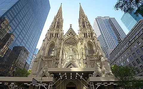 St. Patrick's Cathedral in New York City, a Gothic Revival church, viewed from below, surrounded by modern skyscrapers.