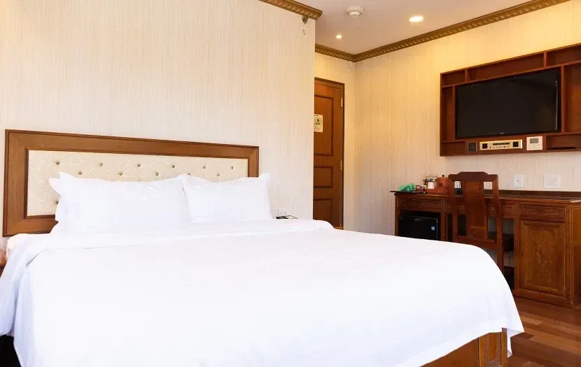 Hotel room with a large bed, TV, desk, and wooden furniture. White bedding against beige walls.