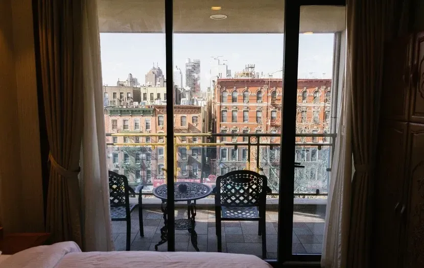 Balcony view: two chairs and table overlook buildings and sky. Curtains frame a window.