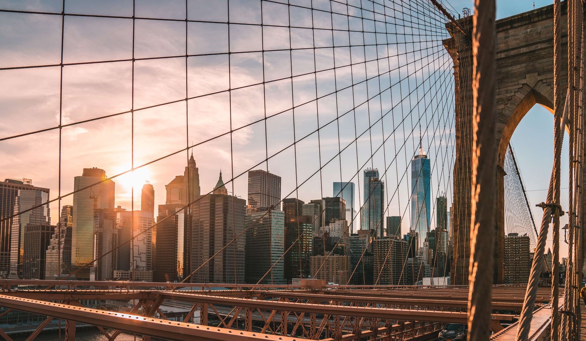 Sunset over NYC skyline viewed through the Brooklyn Bridge cables and arches.