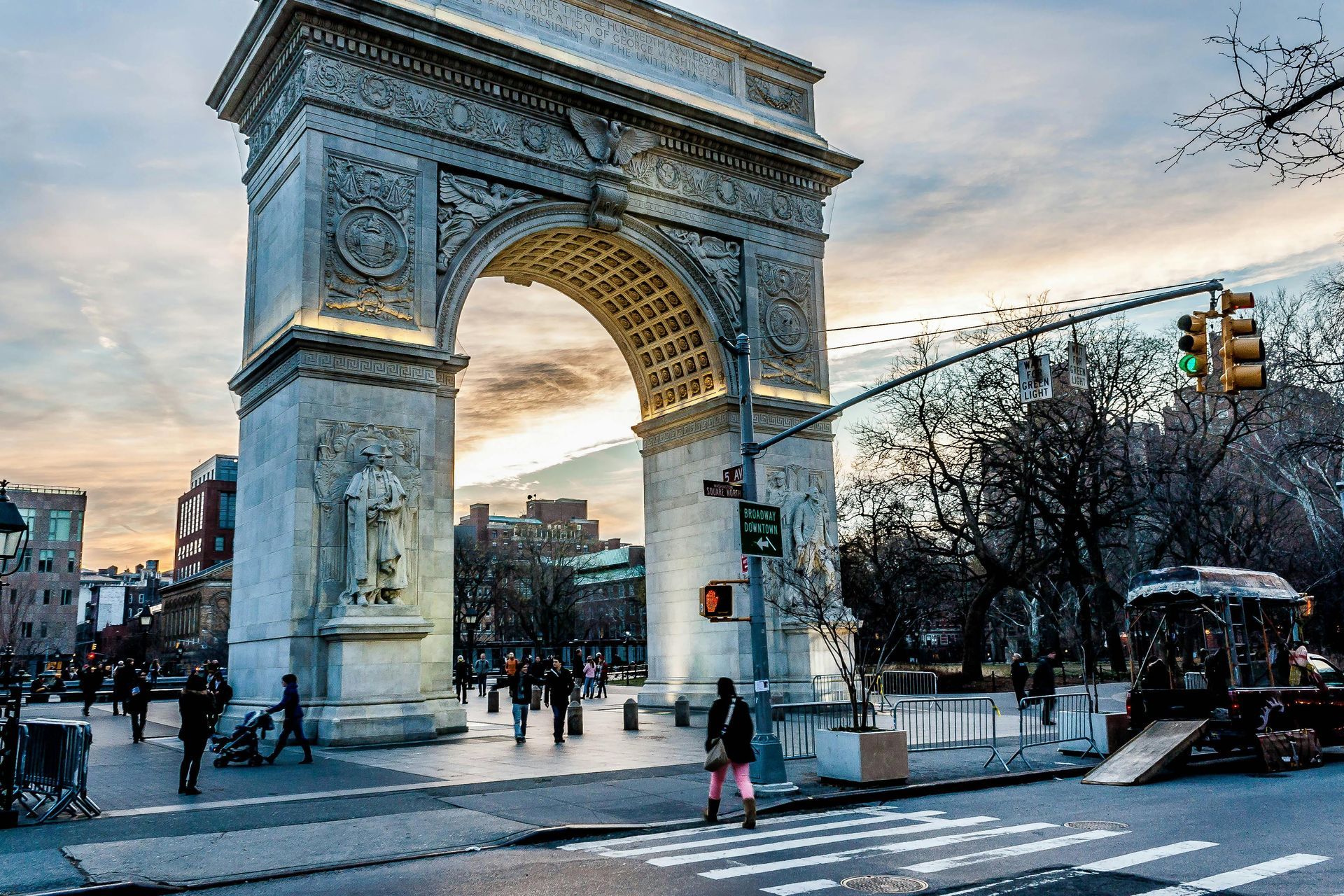 Washington Square Arch, NYC; pedestrians and vehicles on a city street, cloudy sky.