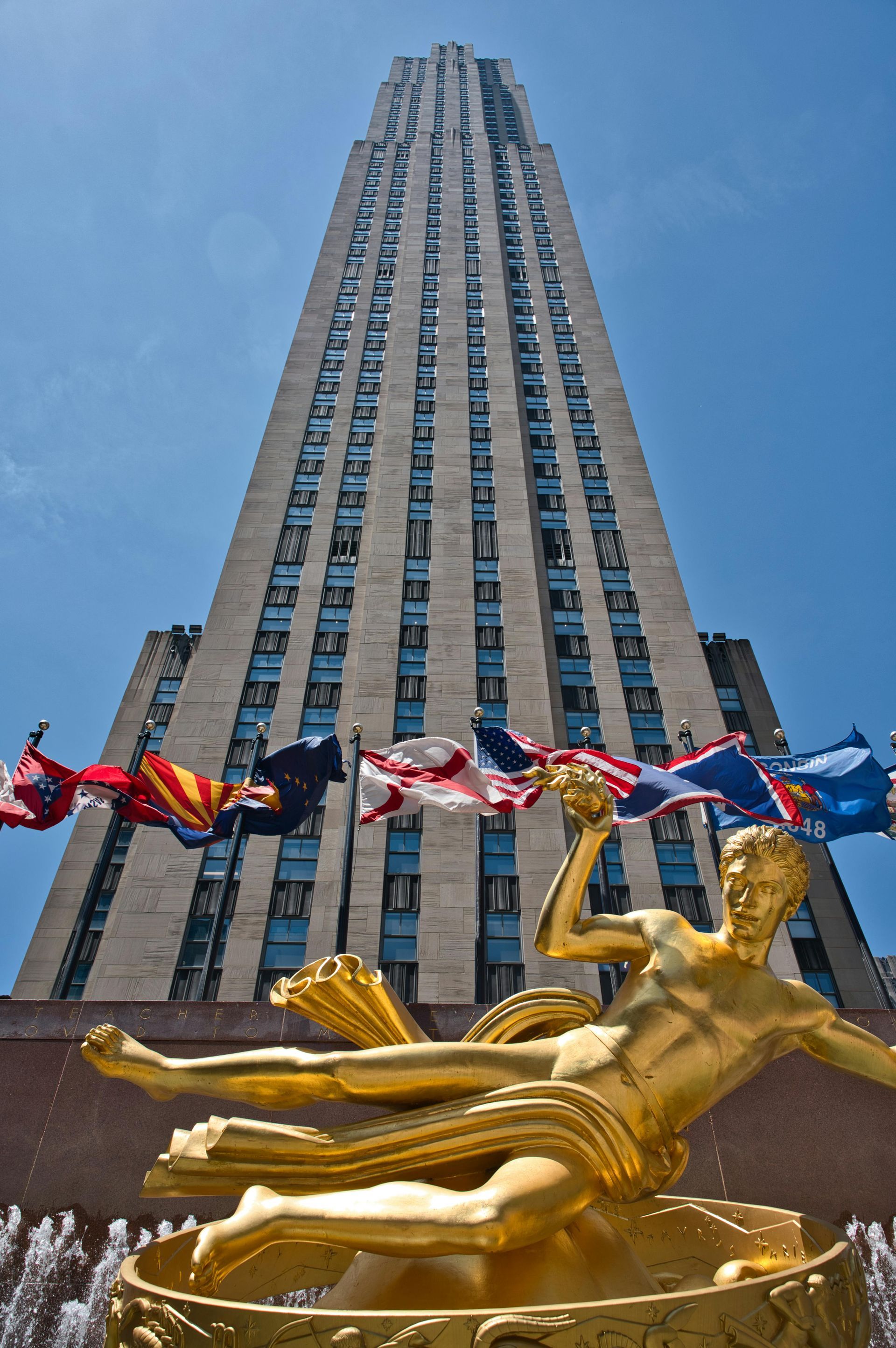 Golden statue and fountain in front of Rockefeller Center skyscraper with flags waving in the sun.