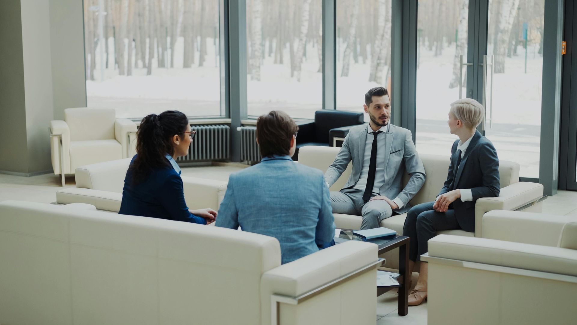 Four people in business attire sit in a bright lounge, talking in a small circle.