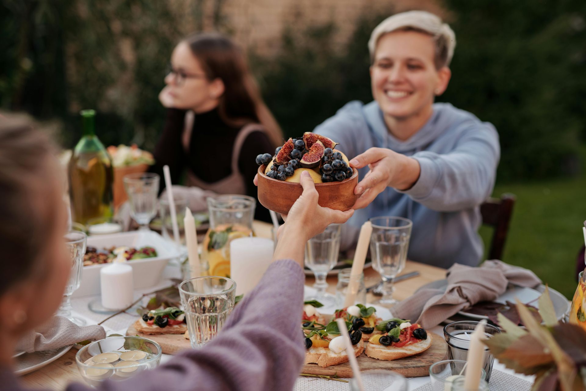 Friends sharing food at an outdoor table, with drinks, plates, and a smiling person offering a muffin