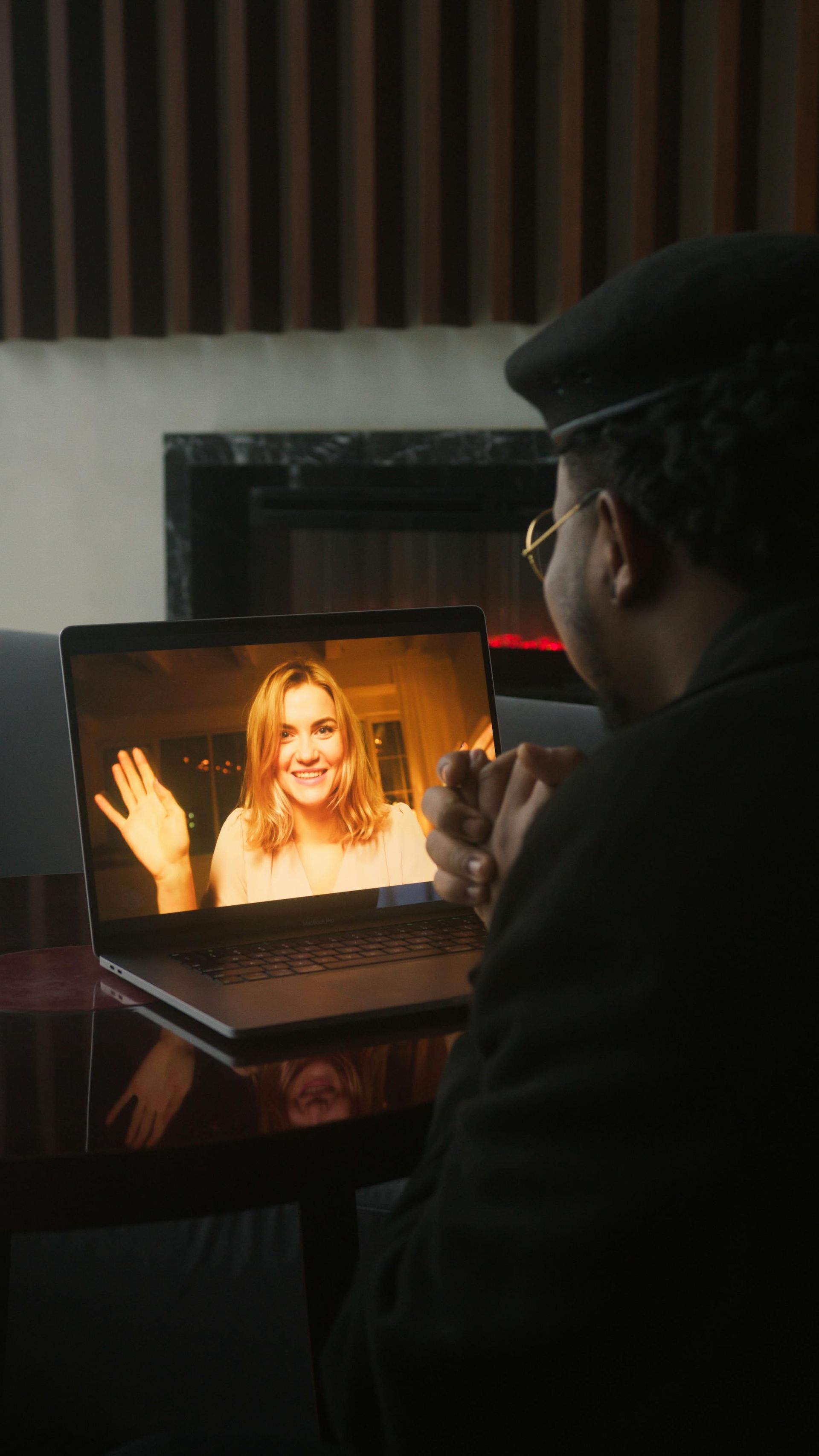 Person in dark hat watching a laptop video call with a smiling woman waving on screen