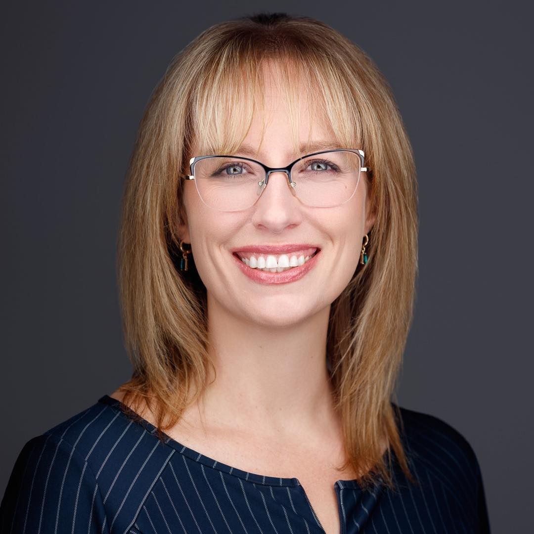 Woman with blonde hair, glasses, and a striped blue top smiles at the camera against a grey backdrop.