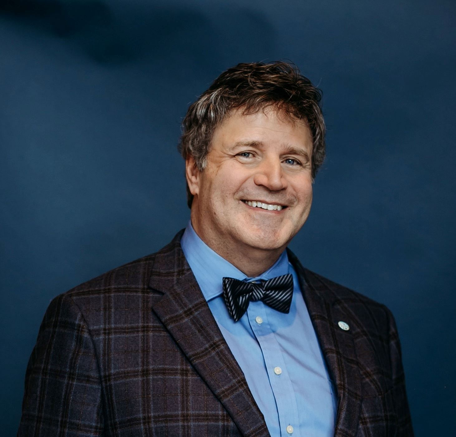 Man in plaid blazer and bow tie, smiling, blue background.