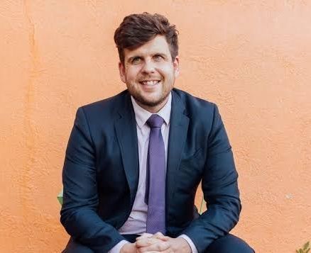 Man in a blue suit and purple tie smiles in front of an orange wall.