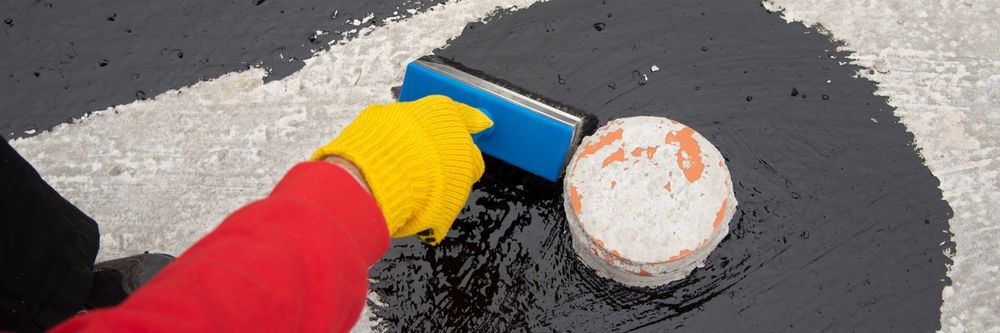 A Person Wearing Gloves is Using a Blue Sponge to Spread Asphalt on the Ground — Legend Waterproofing In New Castle, NSW
