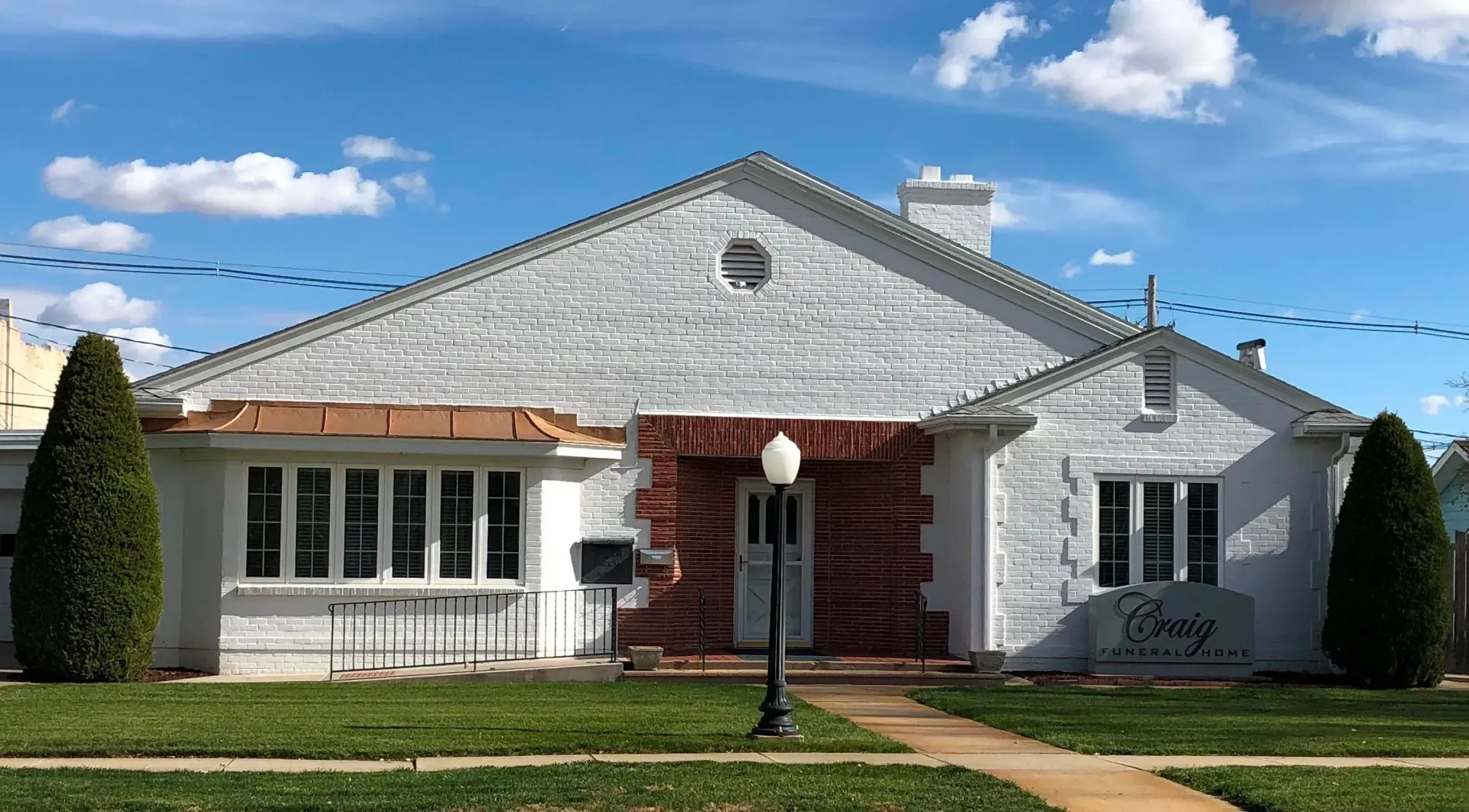White brick building with a gabled roof, a central door, and a sign on the right; blue sky with clouds.