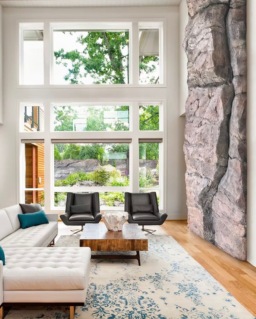 Living room with large windows overlooking trees, white sectional, black chairs, and stone accent wall.