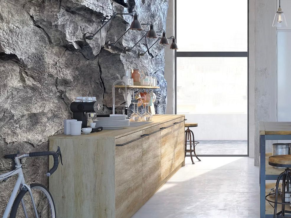 Kitchen with rough stone wall, wooden cabinets, and a bicycle. Sunlight streams through a large window.