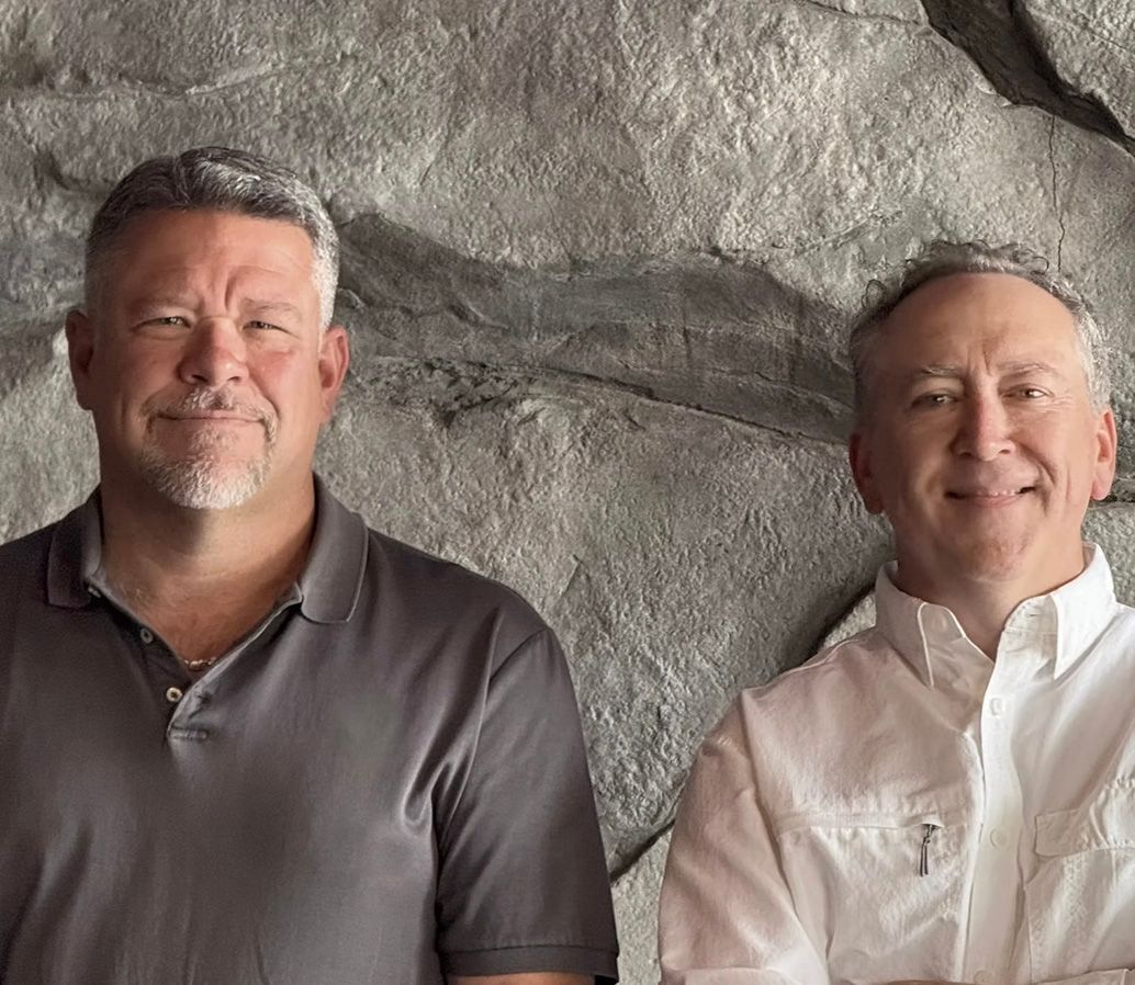 Two men in front of a stone wall. One wears a gray shirt, the other a white shirt, both smiling.