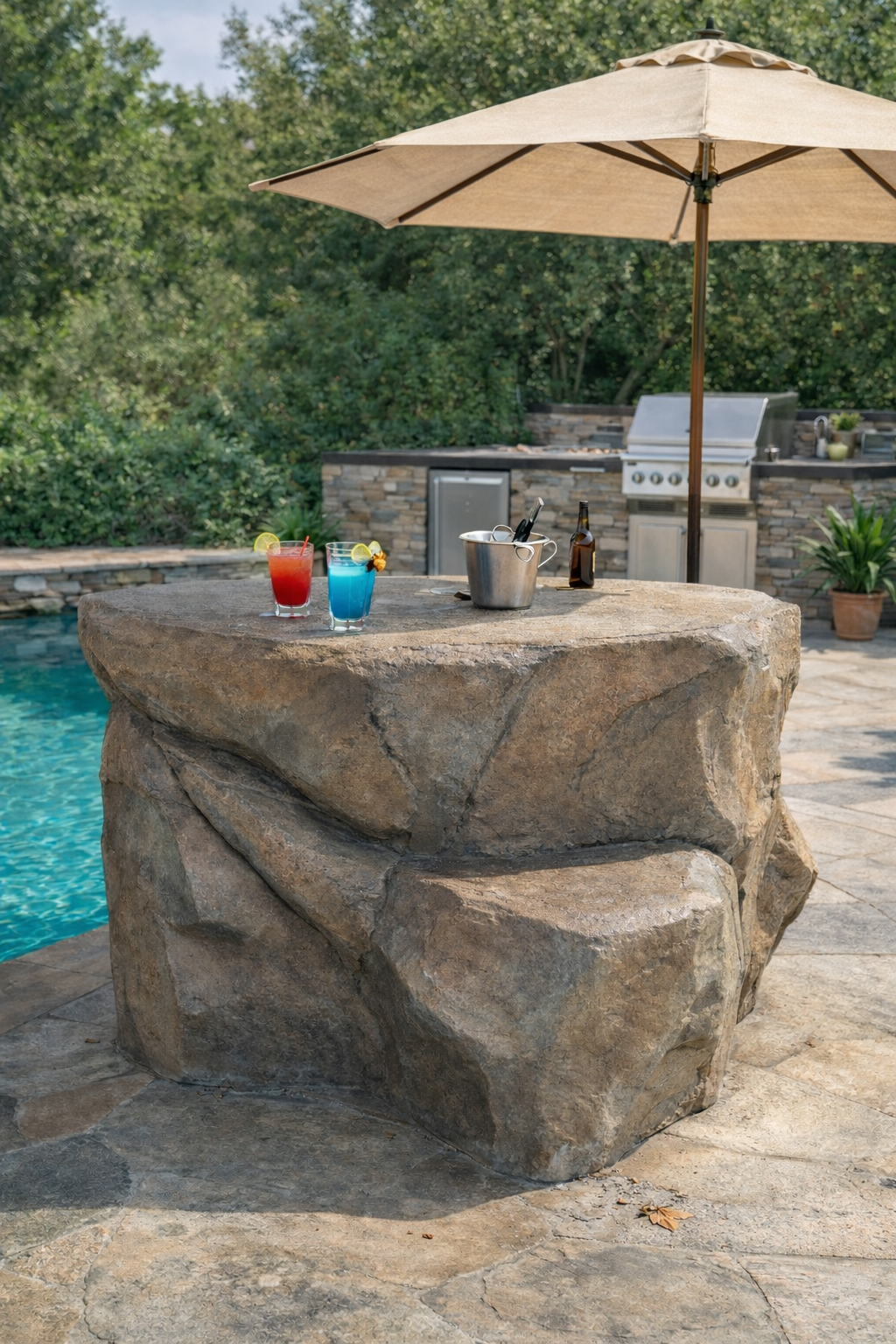 Outdoor bar area beside a pool. Rock-like bar with drinks, umbrella, grill in background.