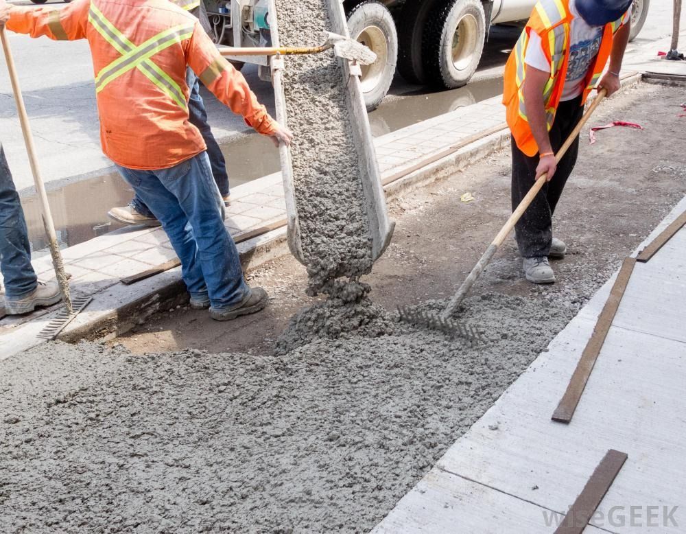 Construction workers in high-visibility vests pour wet concrete from a truck chute onto a sidewalk.