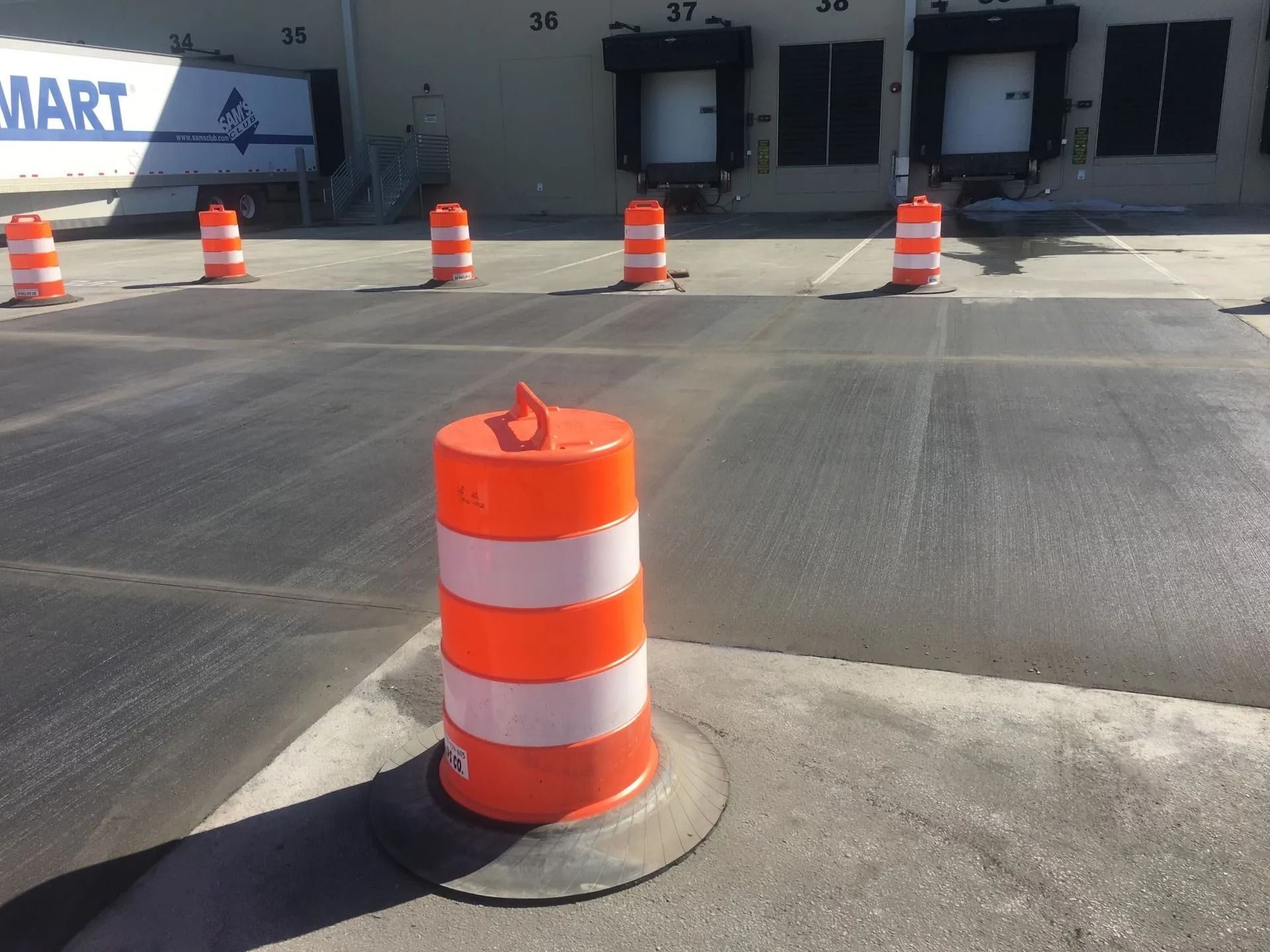 A line of orange and white traffic barrels sits in a warehouse parking lot in front of loading docks.