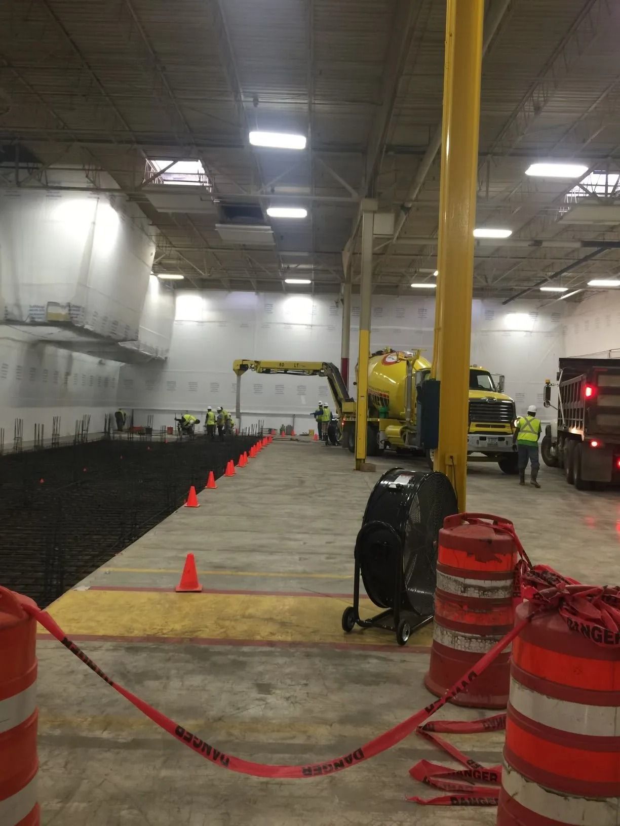 Construction workers in a large, dimly lit warehouse with a yellow concrete truck and safety cones on a concrete floor.