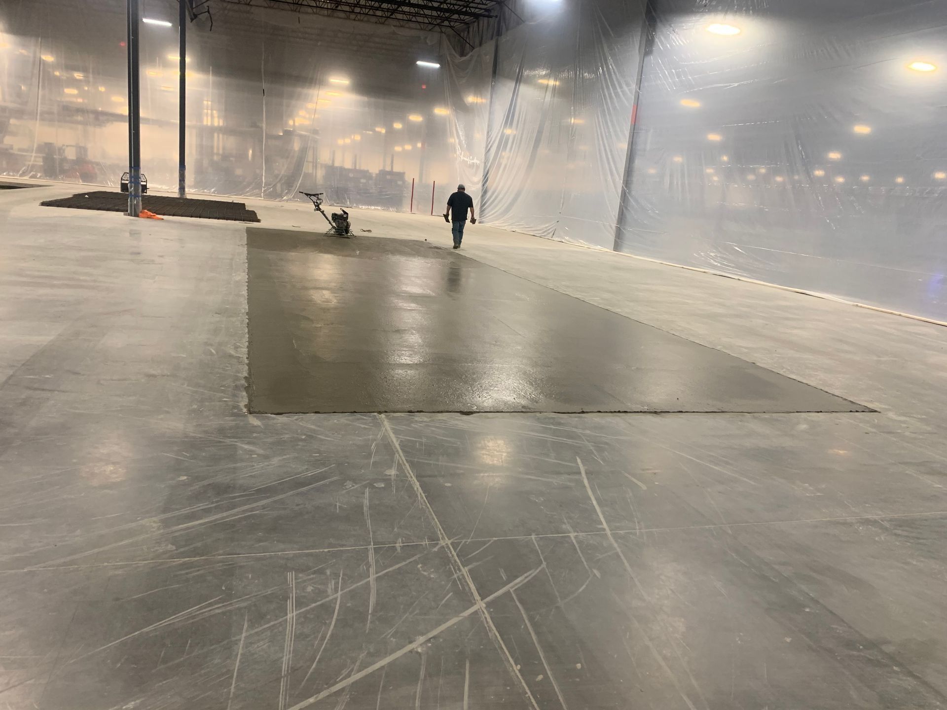 A construction worker operates a power trowel on a rectangular patch of wet concrete inside a large warehouse.