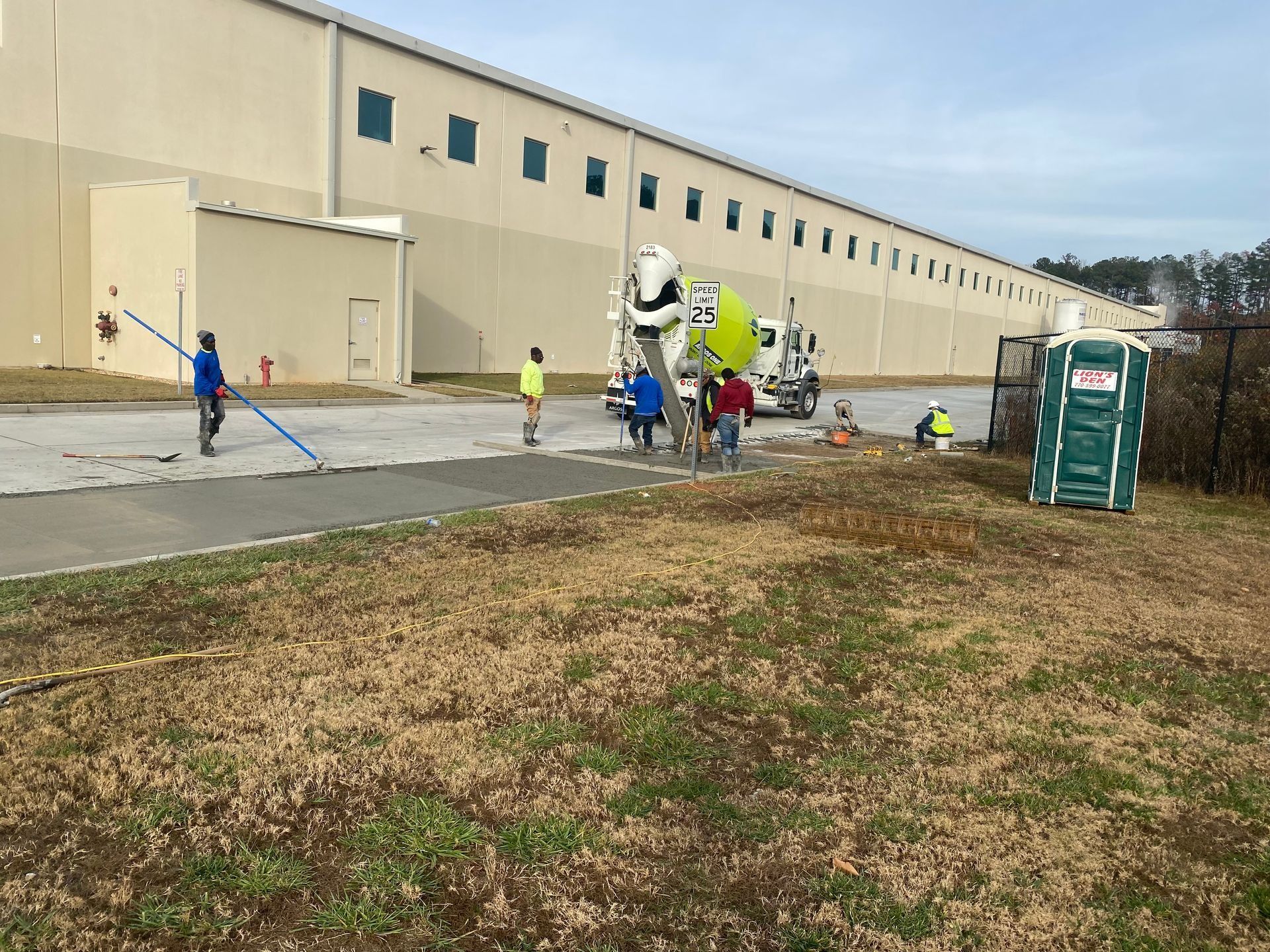 Construction workers prepare concrete for a sidewalk alongside a large warehouse building.