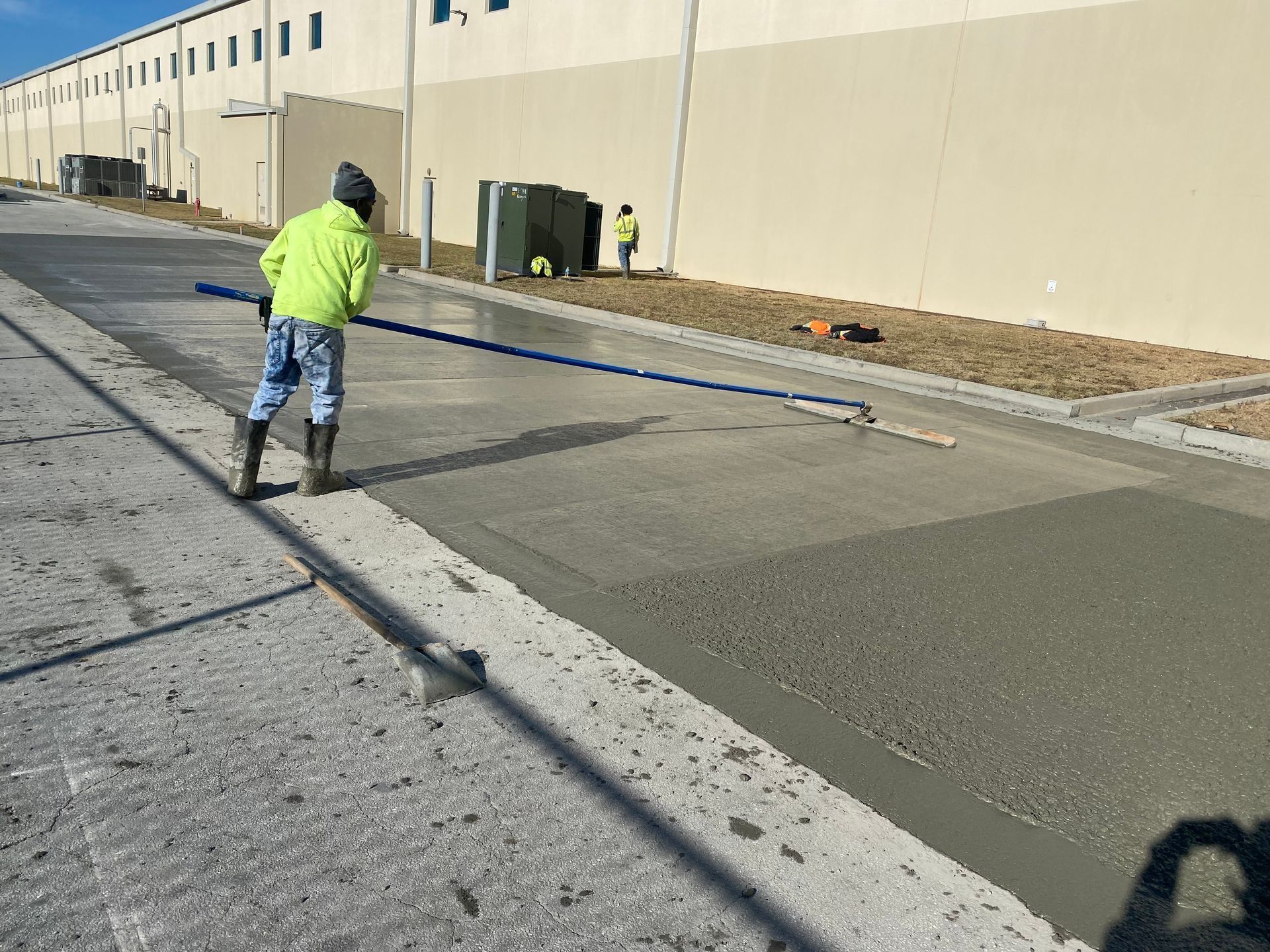 A construction worker in a lime-green jacket uses a long-handled screed to smooth wet concrete next to a large building.