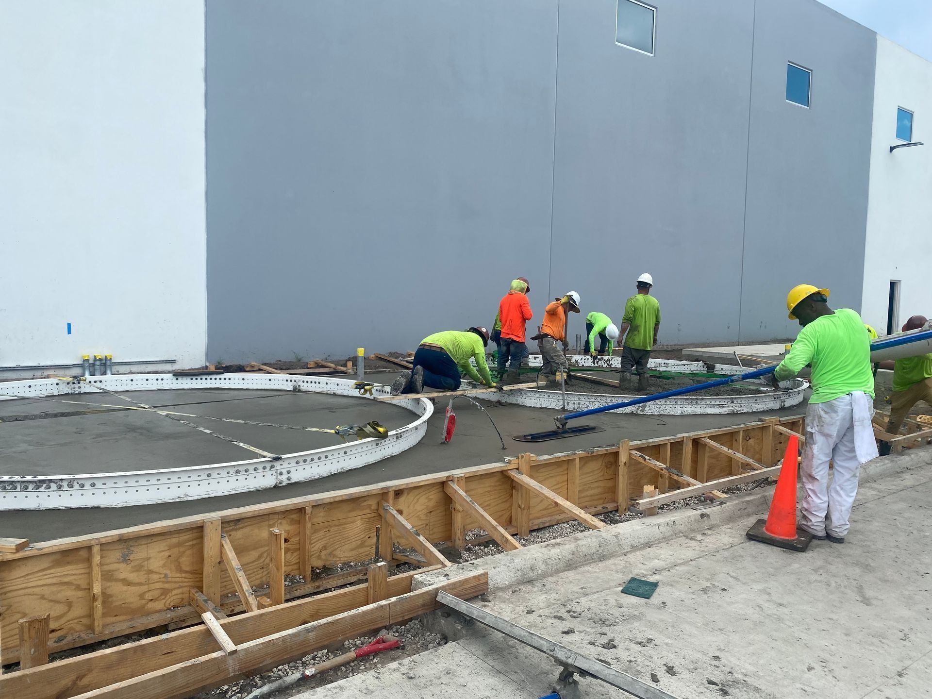 Construction workers smooth fresh concrete into a circular form next to wooden framing outside a warehouse.