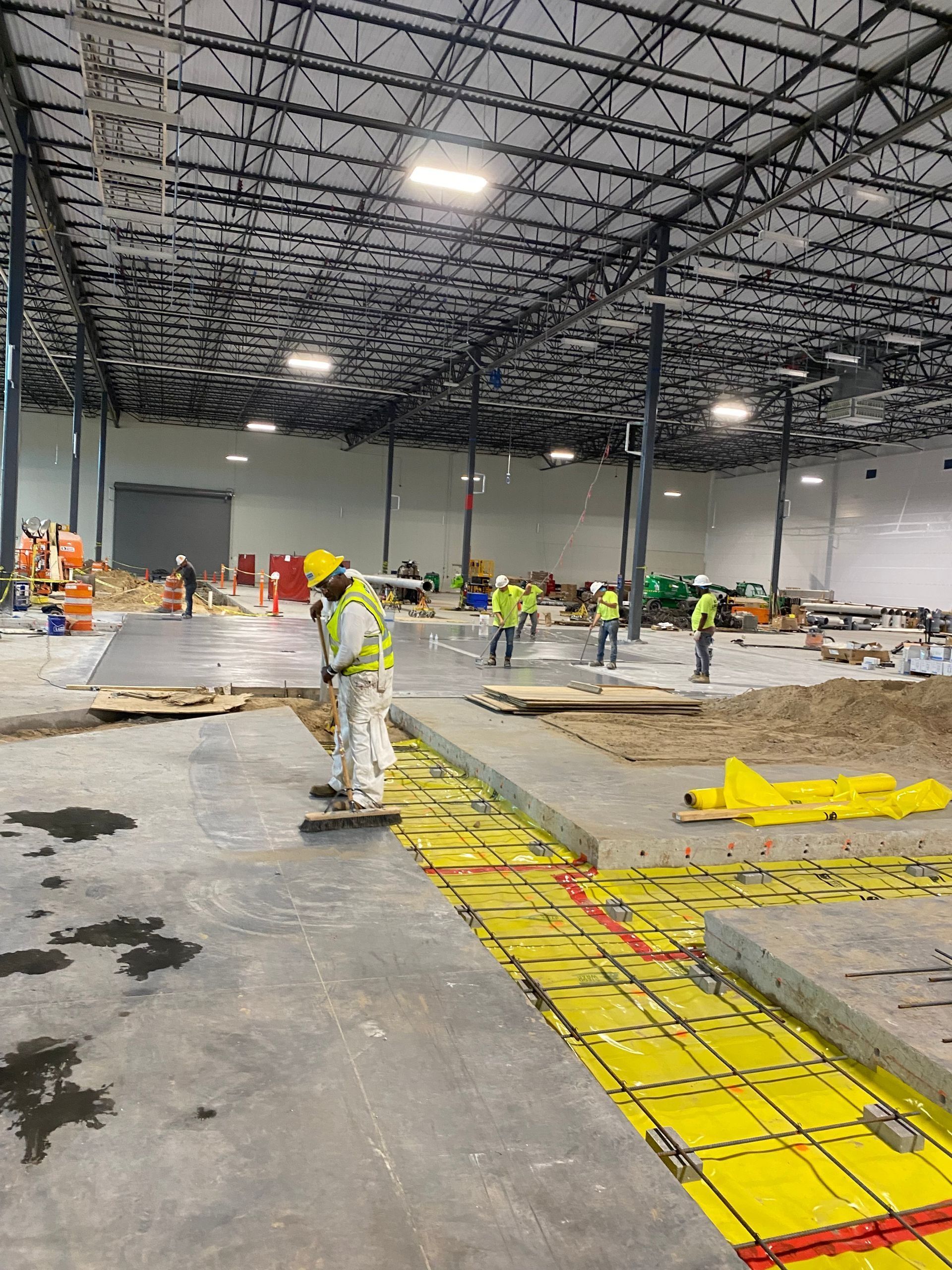 Construction workers wearing high-visibility gear install floor insulation and wiring in a large, unfinished warehouse.