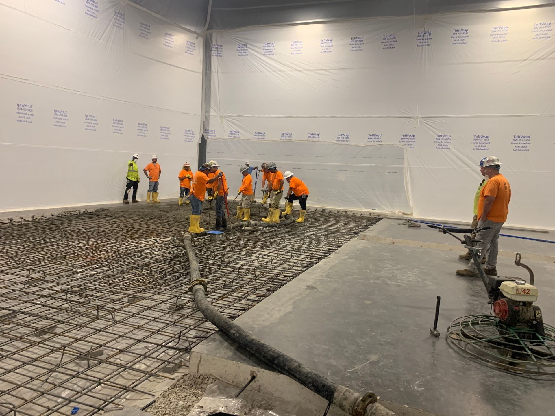 Construction workers in safety gear spreading concrete across a large interior floor, with power trowels nearby.