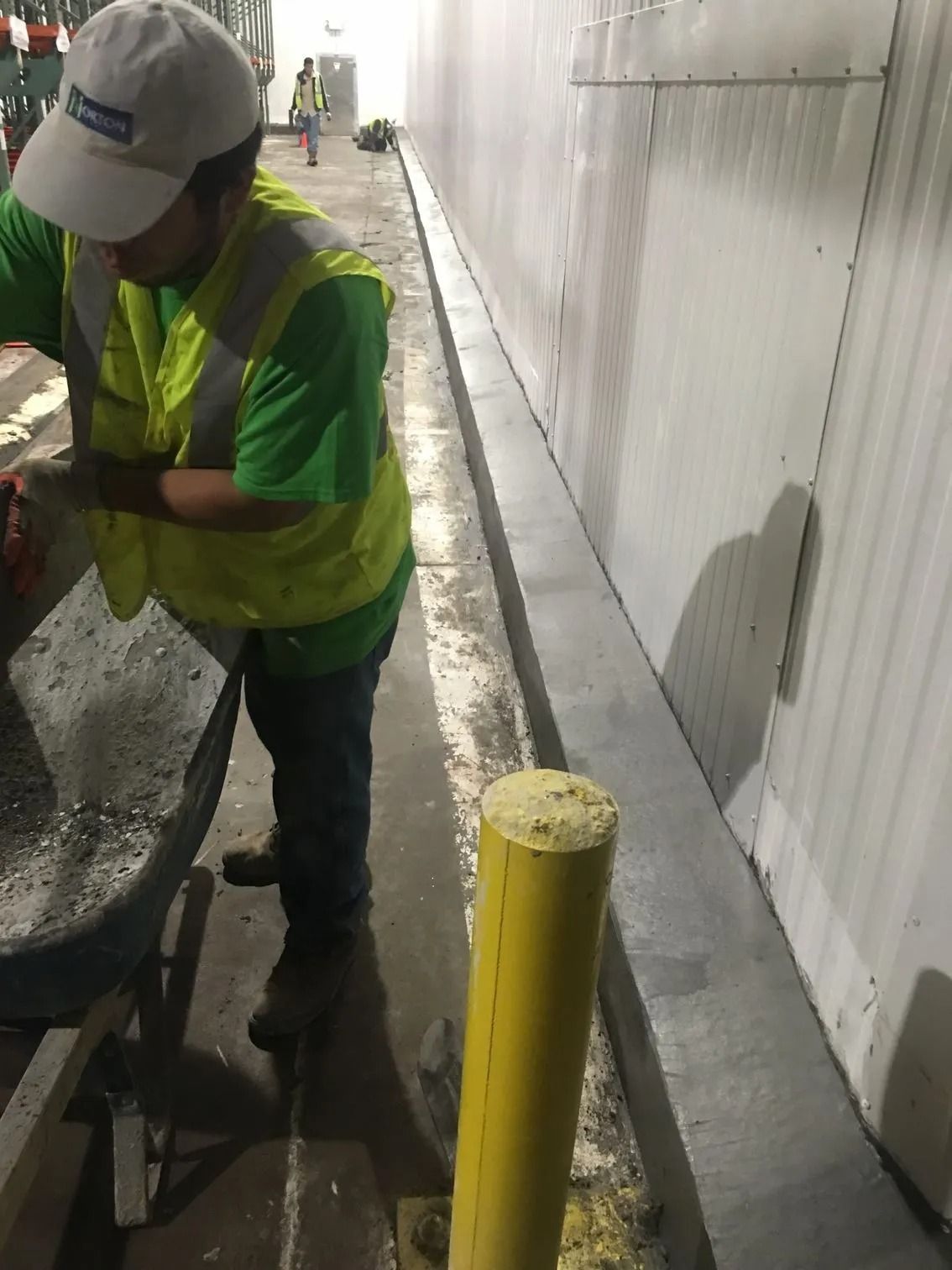 A worker in a high-visibility vest and white cap works with a wheelbarrow near a wall and a yellow protective bollard.