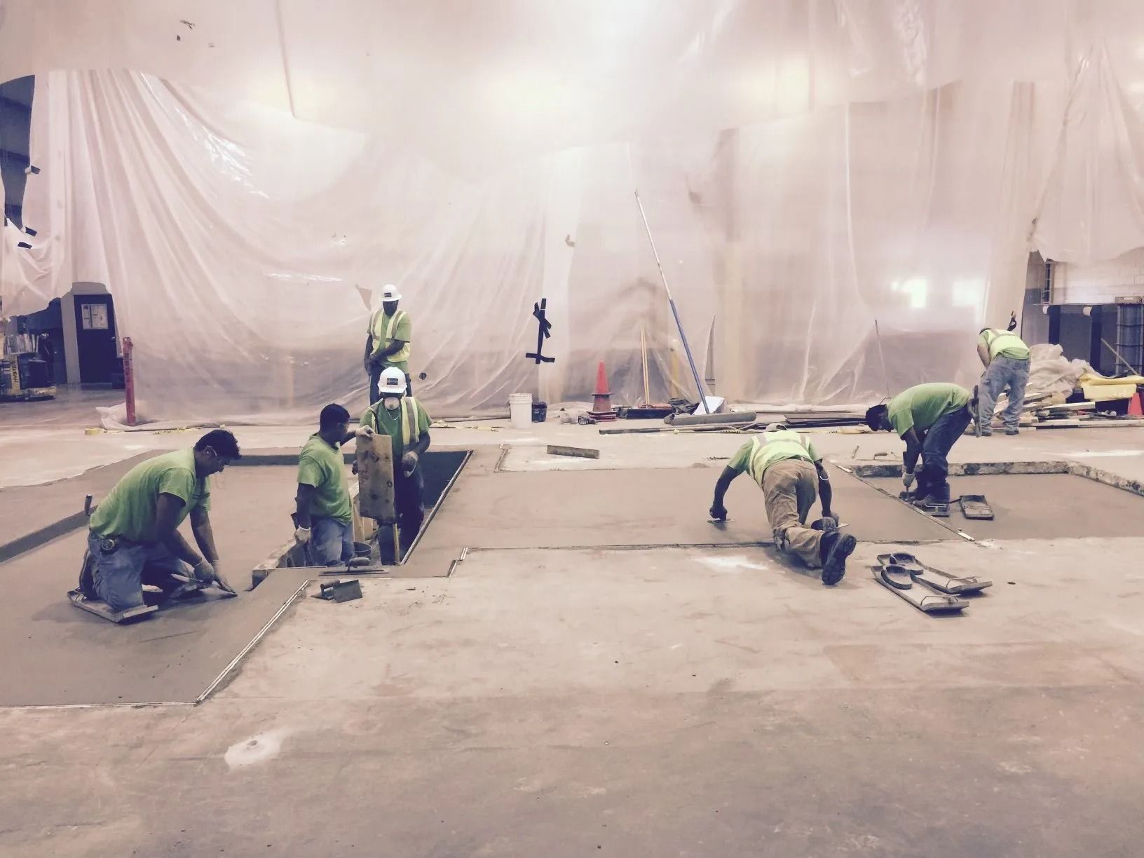 Workers in high-visibility neon shirts smooth freshly poured concrete in an indoor construction site.