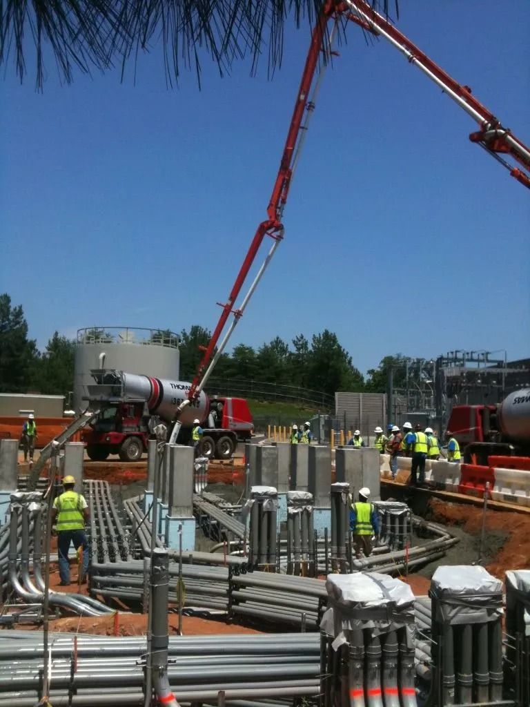 A construction site featuring a red concrete pump truck extending its arm over workers in high-visibility safety vests.
