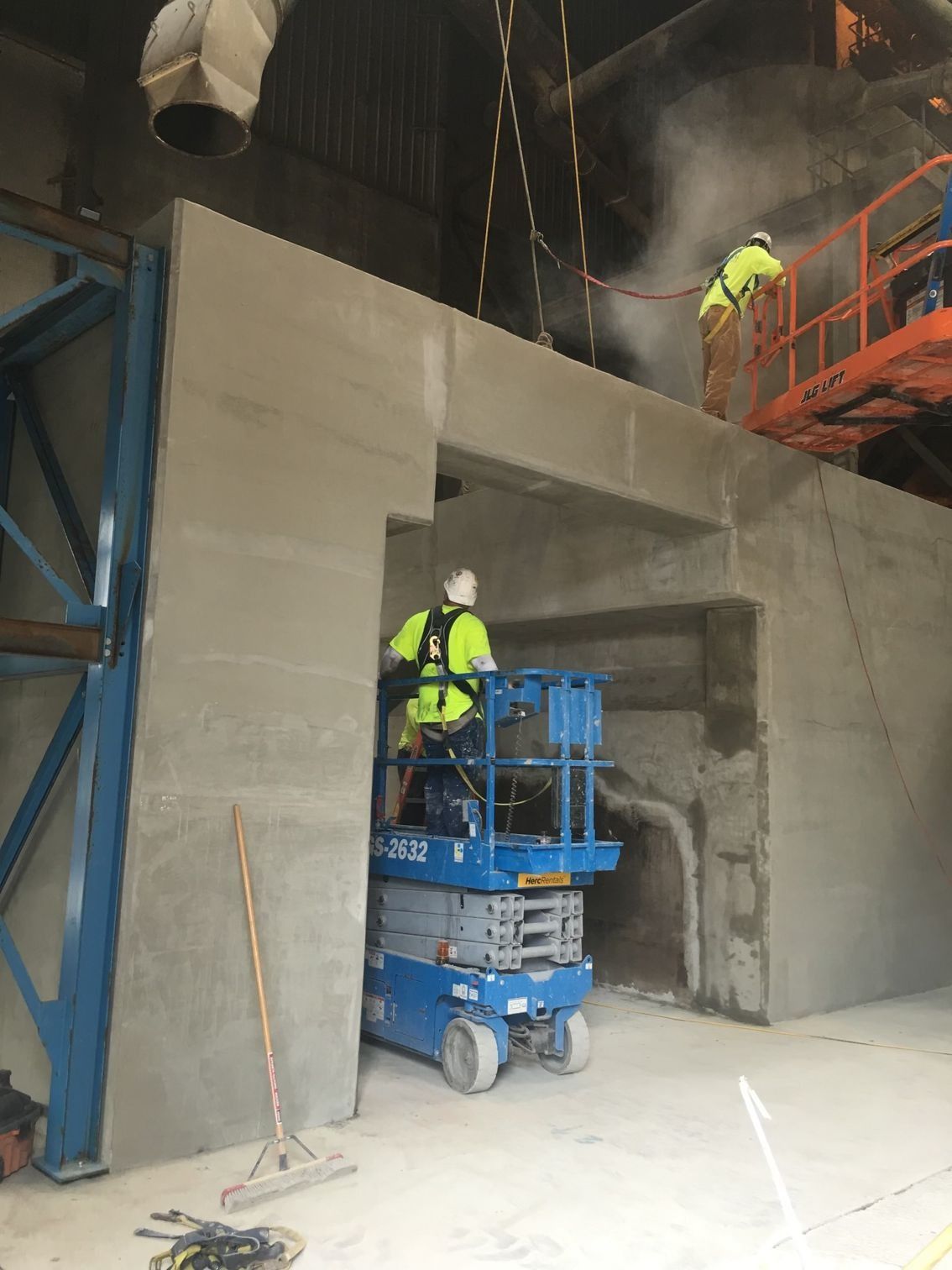 Workers in high-visibility vests use a blue scissor lift to perform maintenance on a large industrial concrete structure.
