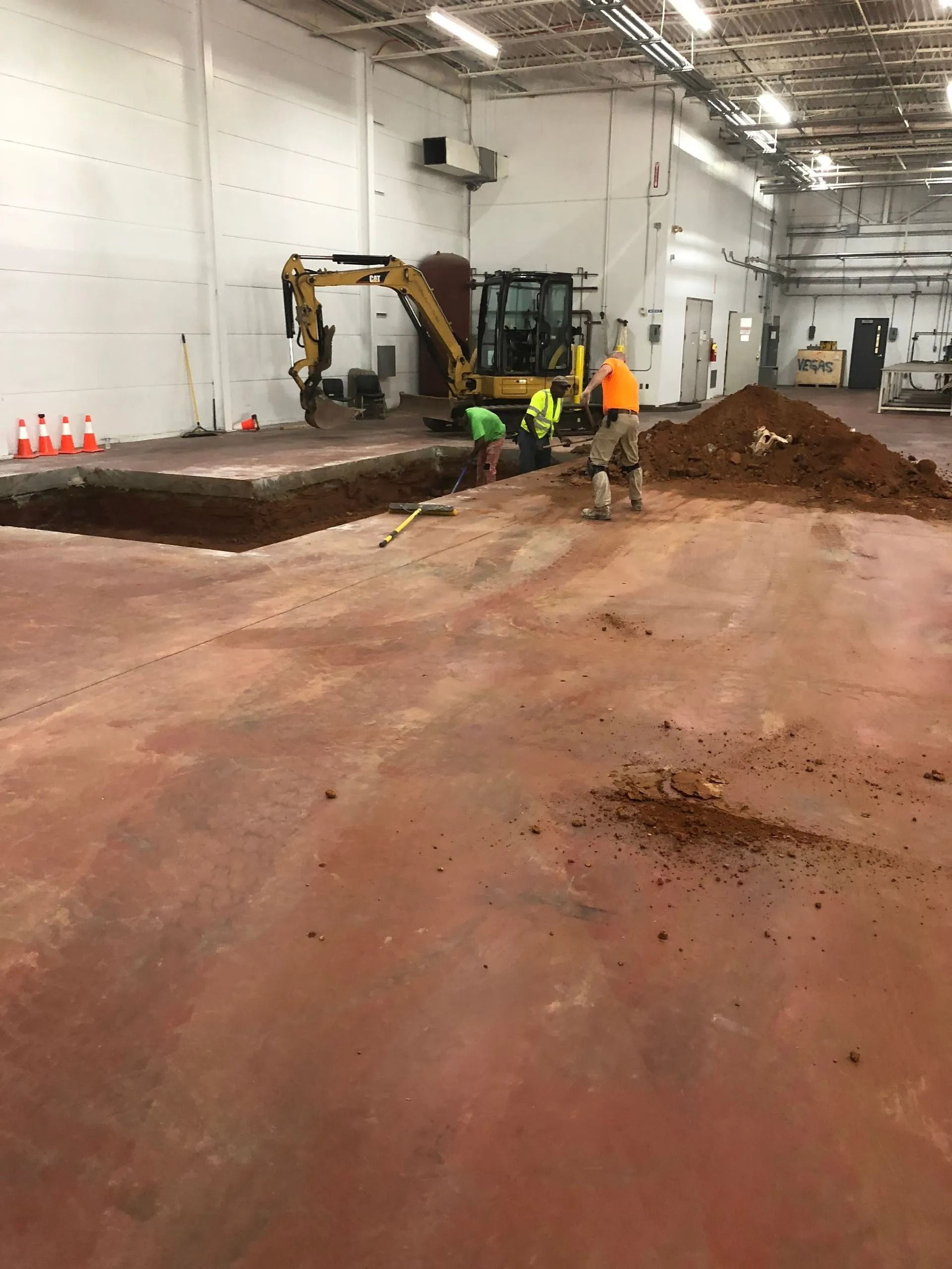 A yellow excavator operates inside a warehouse, digging a trench in the concrete floor while workers stand nearby.