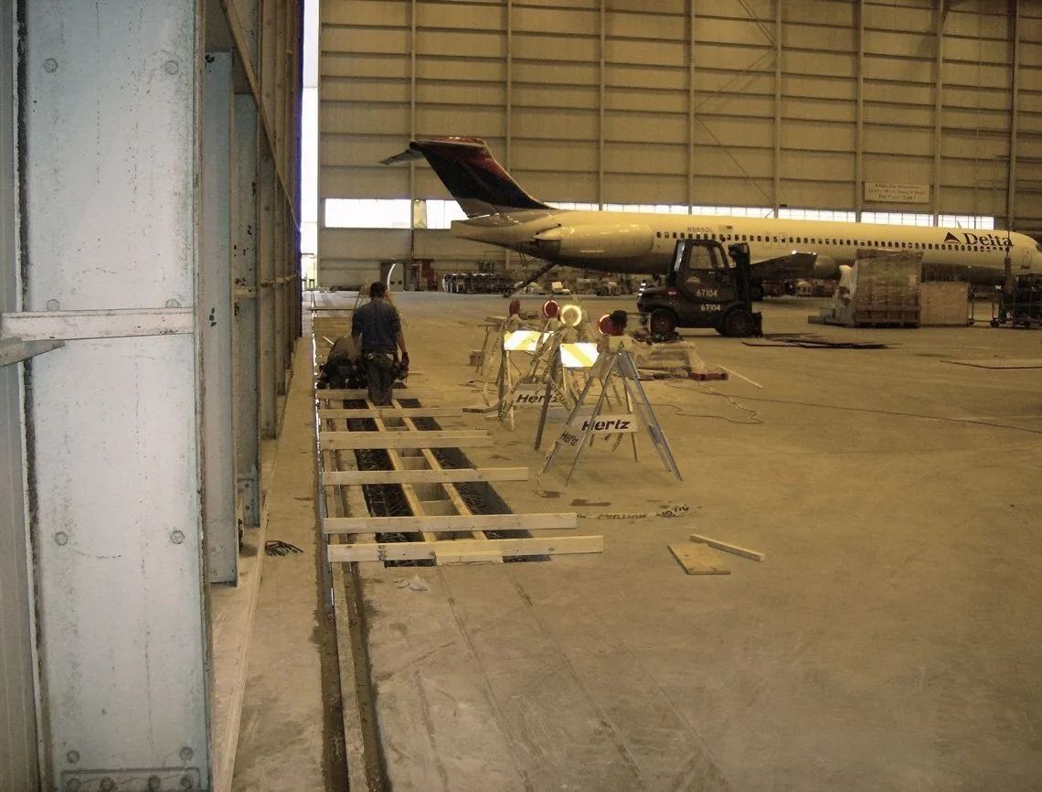 A person works near scaffolding inside an aircraft hangar with a commercial airplane parked in the background.