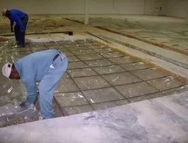 Two workers install a grid of rebar over a plastic vapor barrier on a concrete construction site floor.