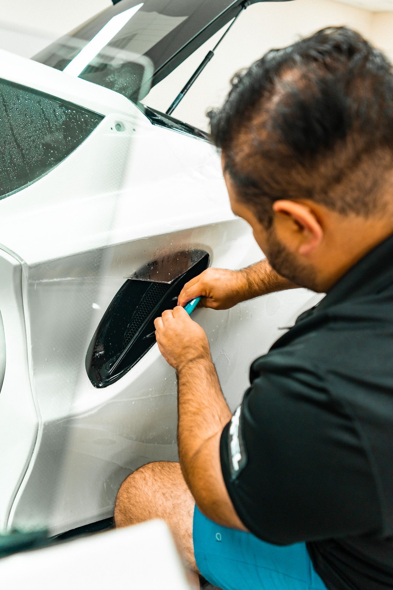 Person spraying a white car panel with a blue bottle, possibly applying a protective film. Red door open.