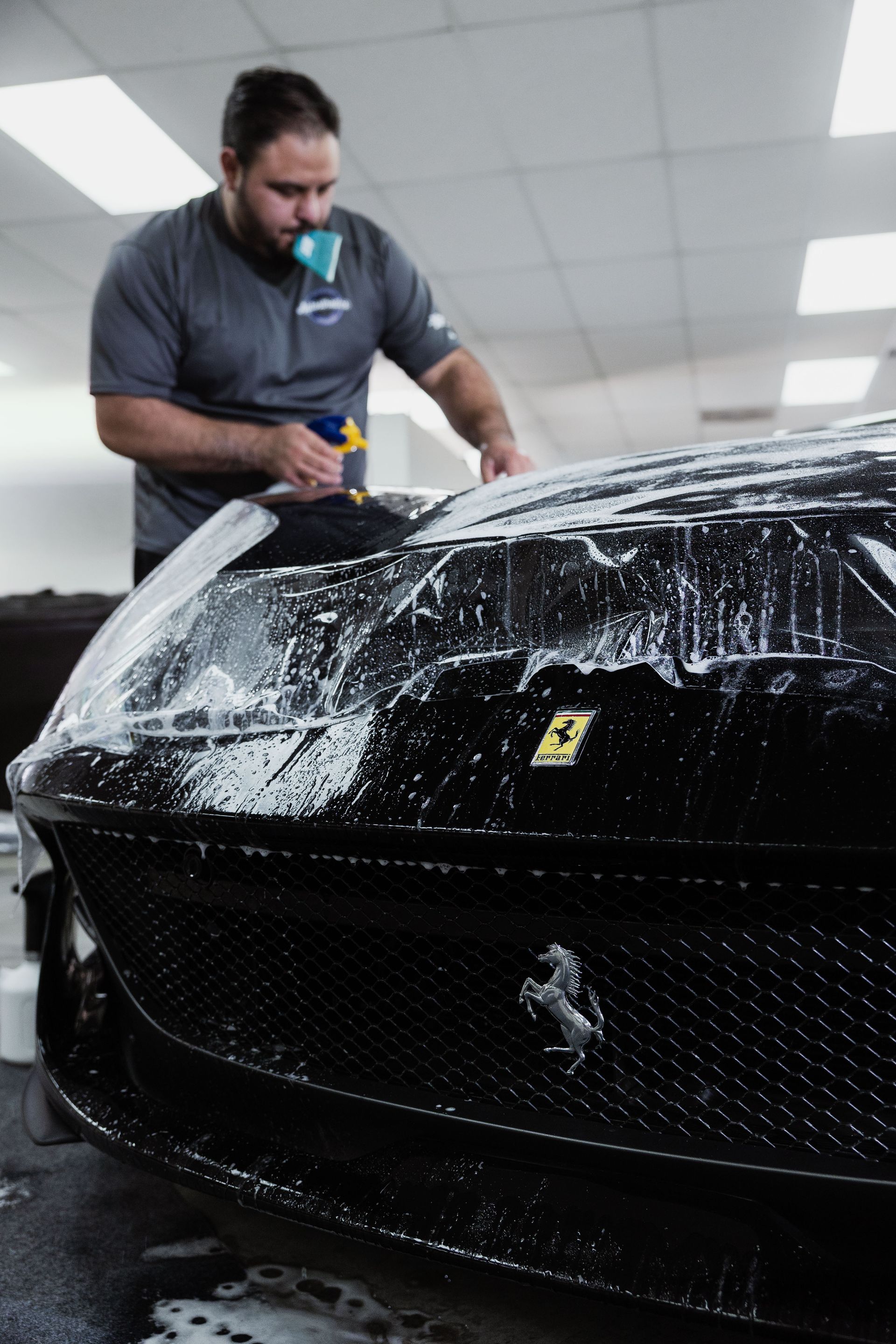 Man sprays soapy water on a black Ferrari in a car detailing shop.