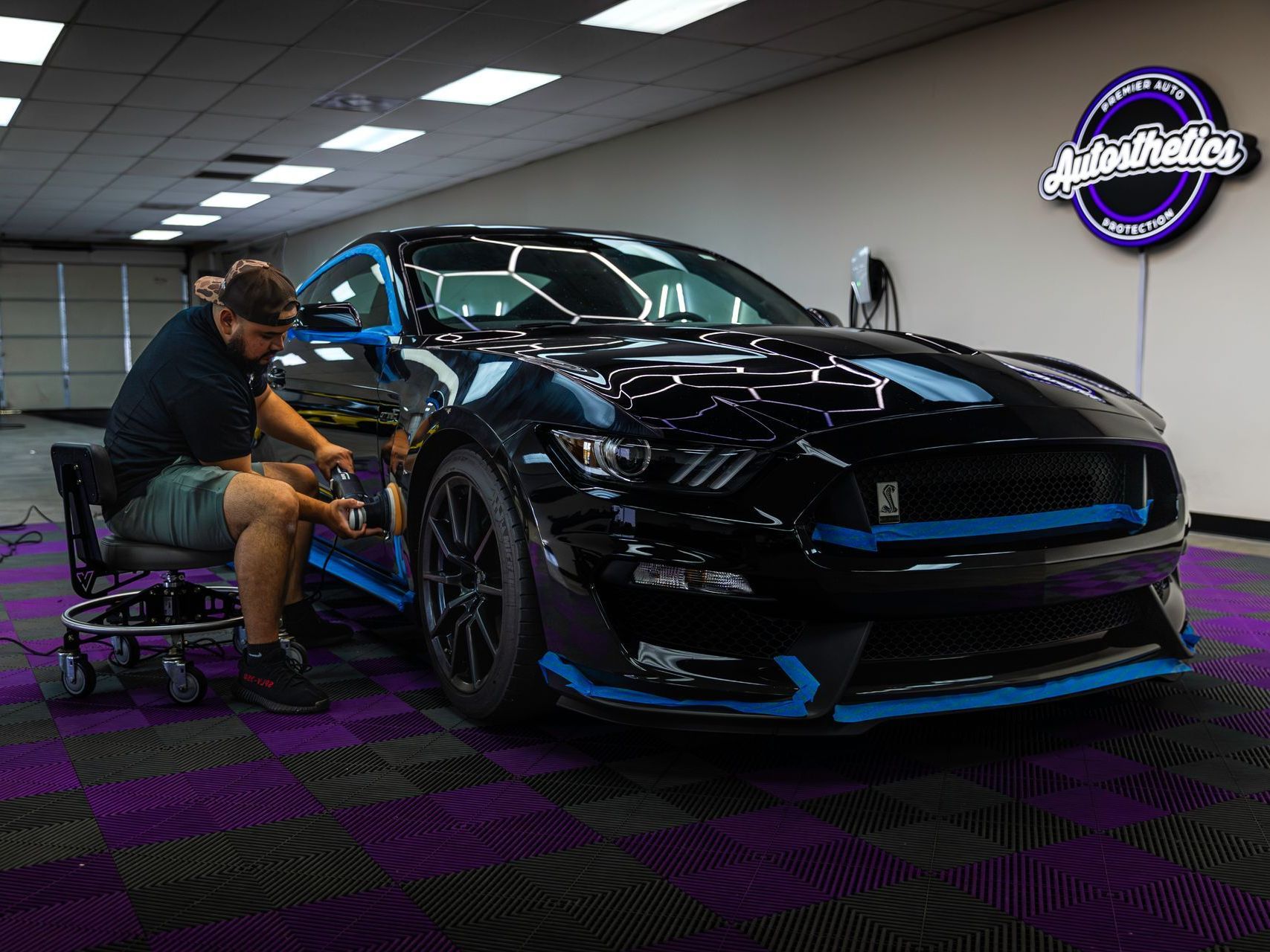 A man is sitting on a stool next to a black and blue mustang in a garage.