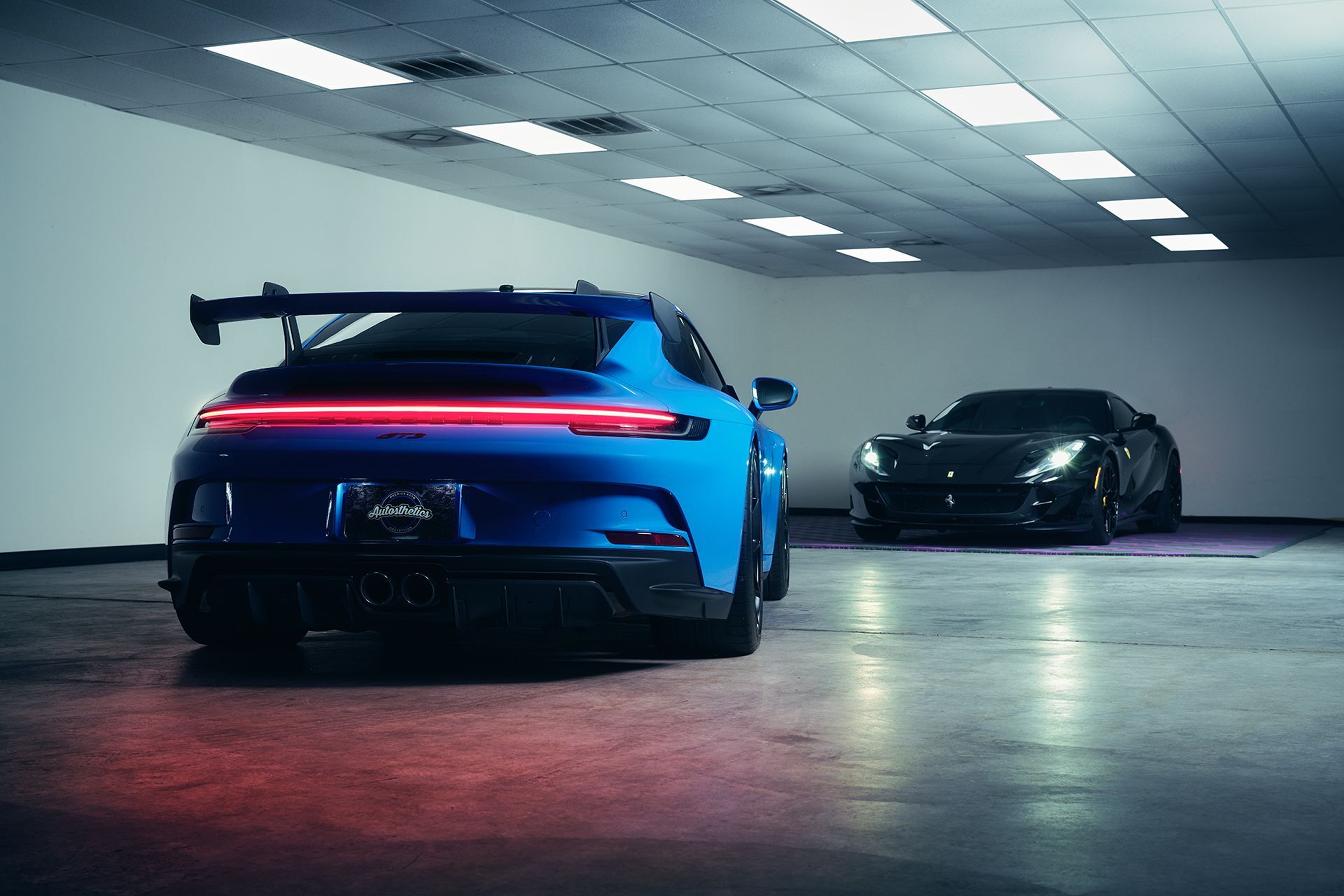 Blue sports car with rear spoiler in a garage, black car in background.