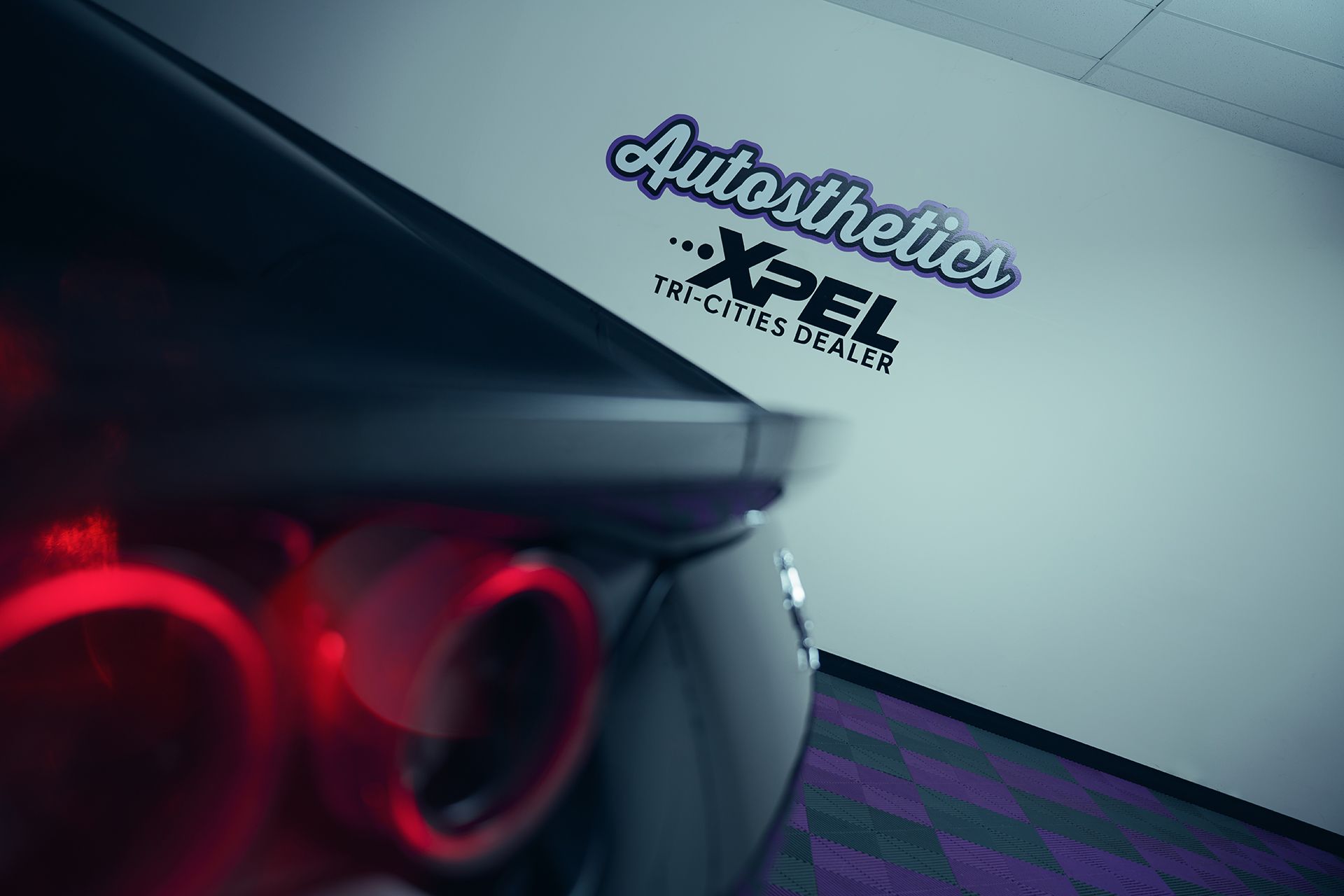 Blurred view of a dark car's rear with red tail lights, near a wall with 