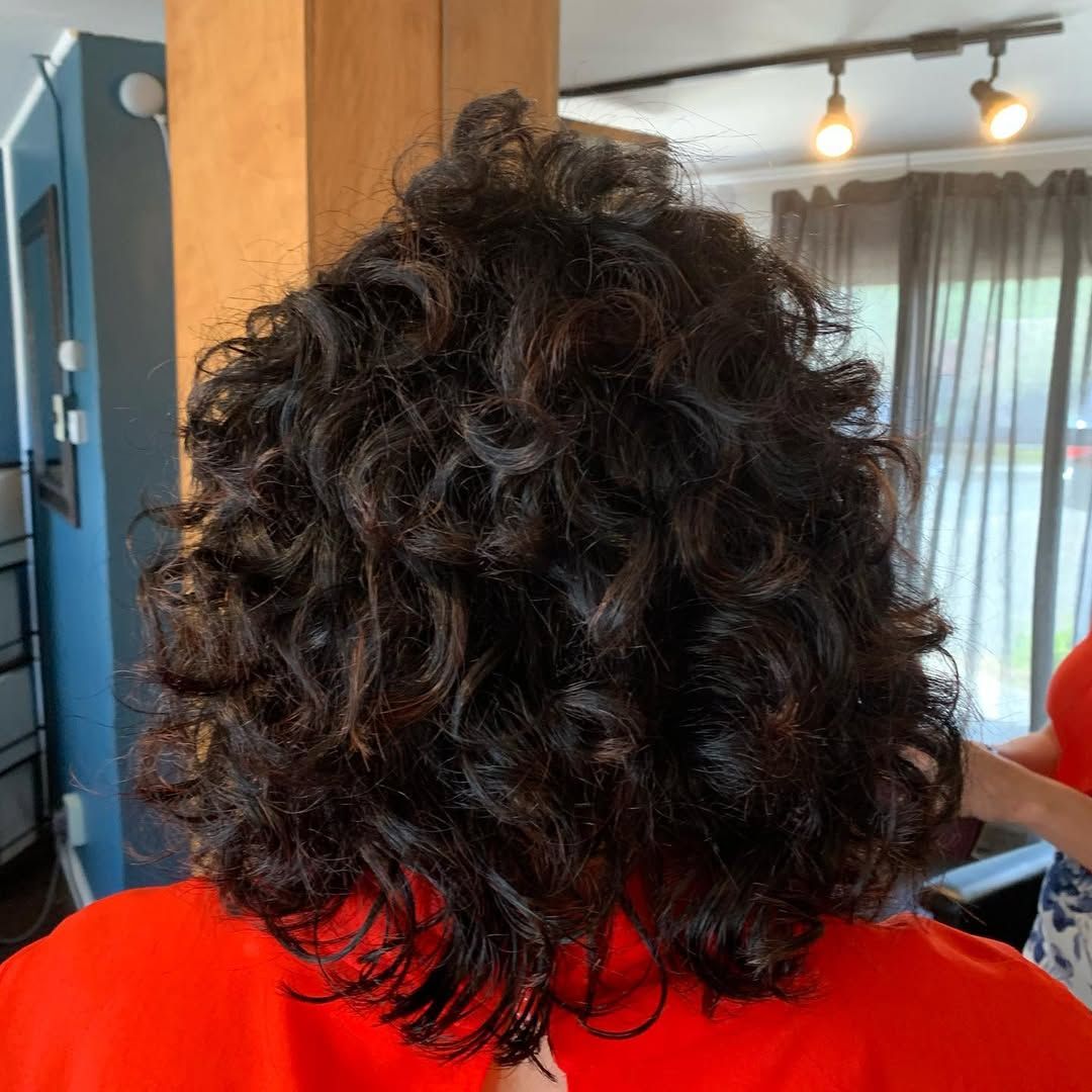 Person with dark, curly hair in a salon chair, facing away. They wear a red shirt.
