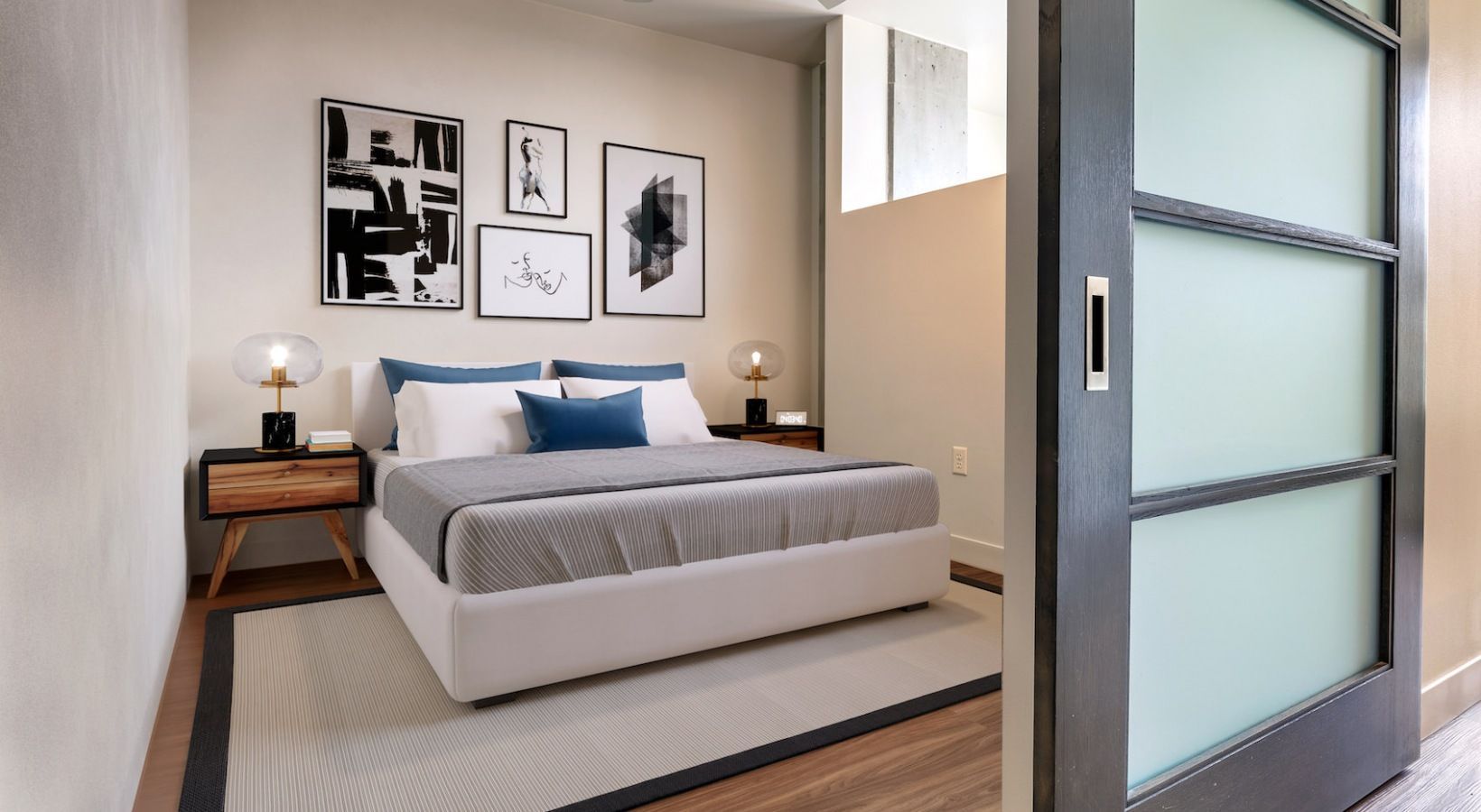 Modern bedroom with a white bed, blue pillows, two nightstands, framed art, and a frosted glass door.