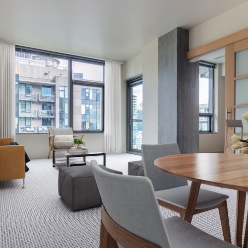 Modern apartment living room with seating, a round wooden table, and large windows.