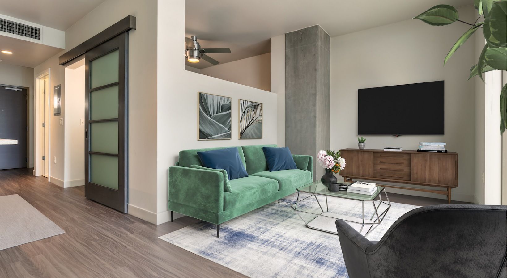 Interior apartment living room with a green sofa, glass coffee table, and wall-mounted TV.