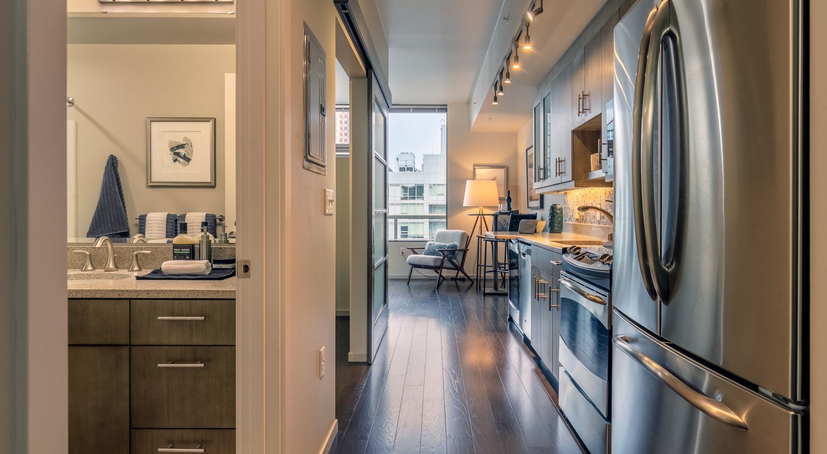 Interior view of a modern apartment: galley kitchen with stainless appliances to the right, bathroom to the left, living area ahead.