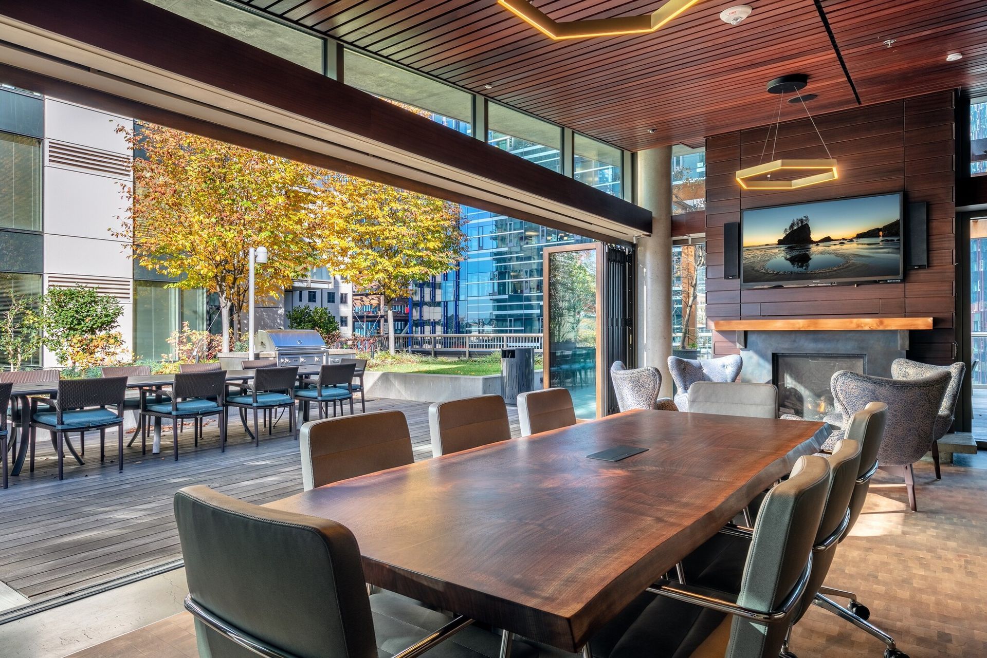 A modern meeting space with a large wooden table, a fireplace, and an outdoor patio with a dining area.