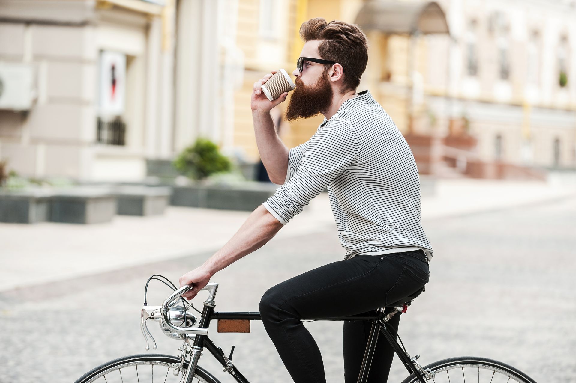 Man with beard drinking from a coffee cup while riding a bicycle on a city street.