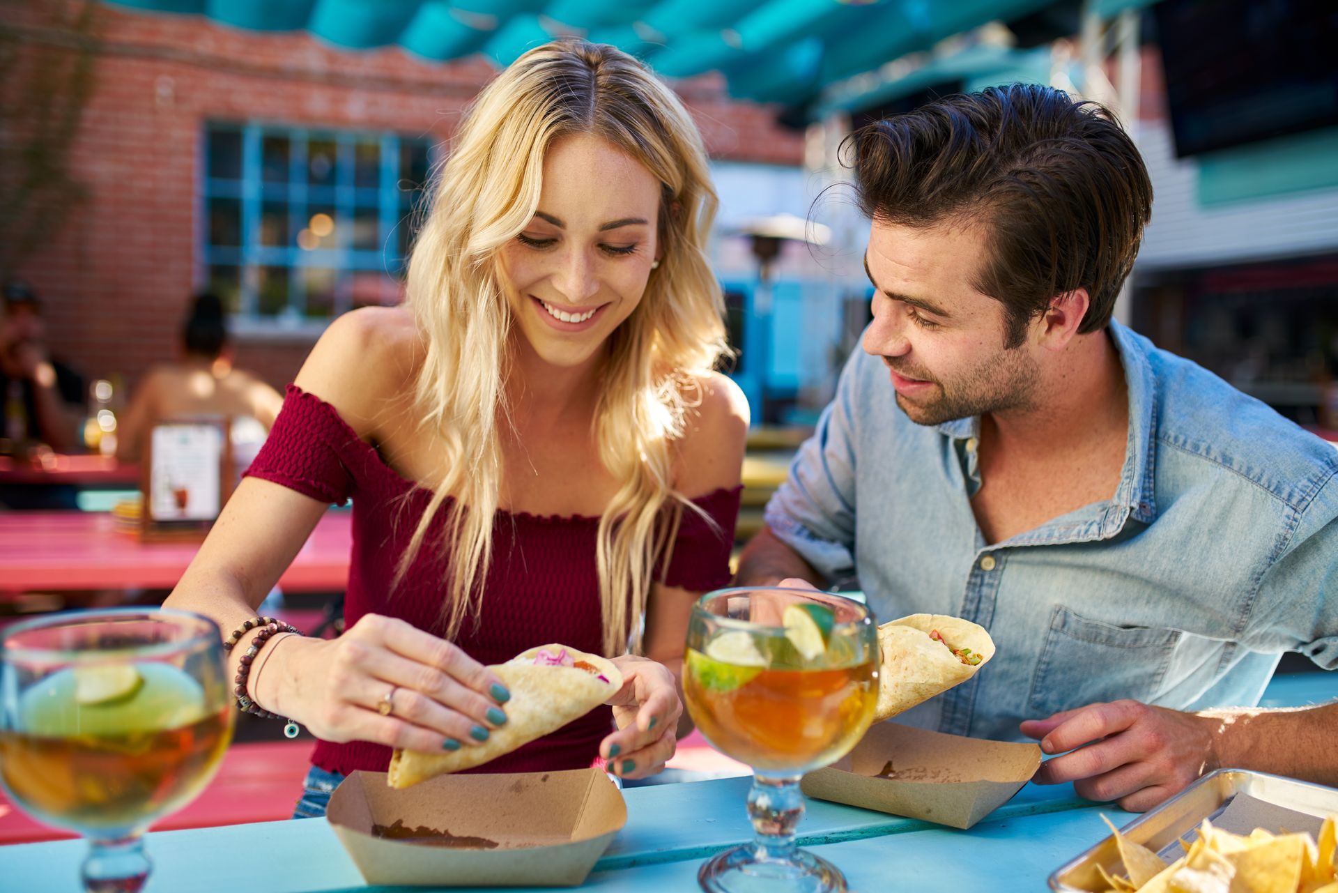 Couple at an outdoor table, smiling while preparing and eating tacos with drinks.