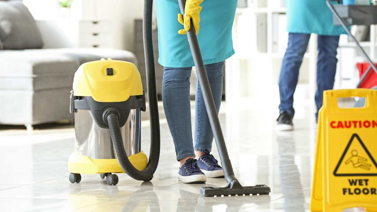 A woman vacuums the floor while a yellow caution sign warns of potential slip hazards nearby.